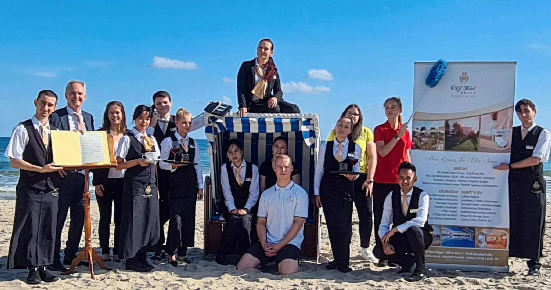 Azubis stehen in Uniformen der Privathotels Dr. Lohbeck am Strand vor dem Meer und lächeln in die Kamera