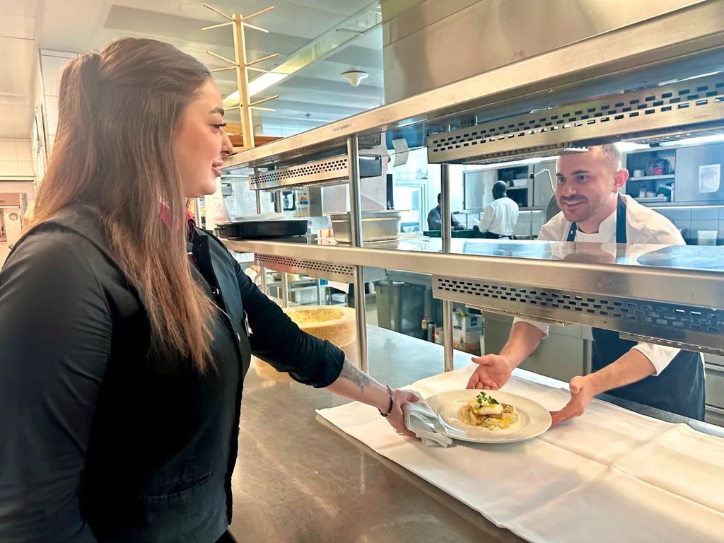 Kochazubi Hayri Akar reicht der angehenden Hotelfachfrau Selina Duman eine frisch zubereitete Hauptspeise f&uuml;r einen Gast.