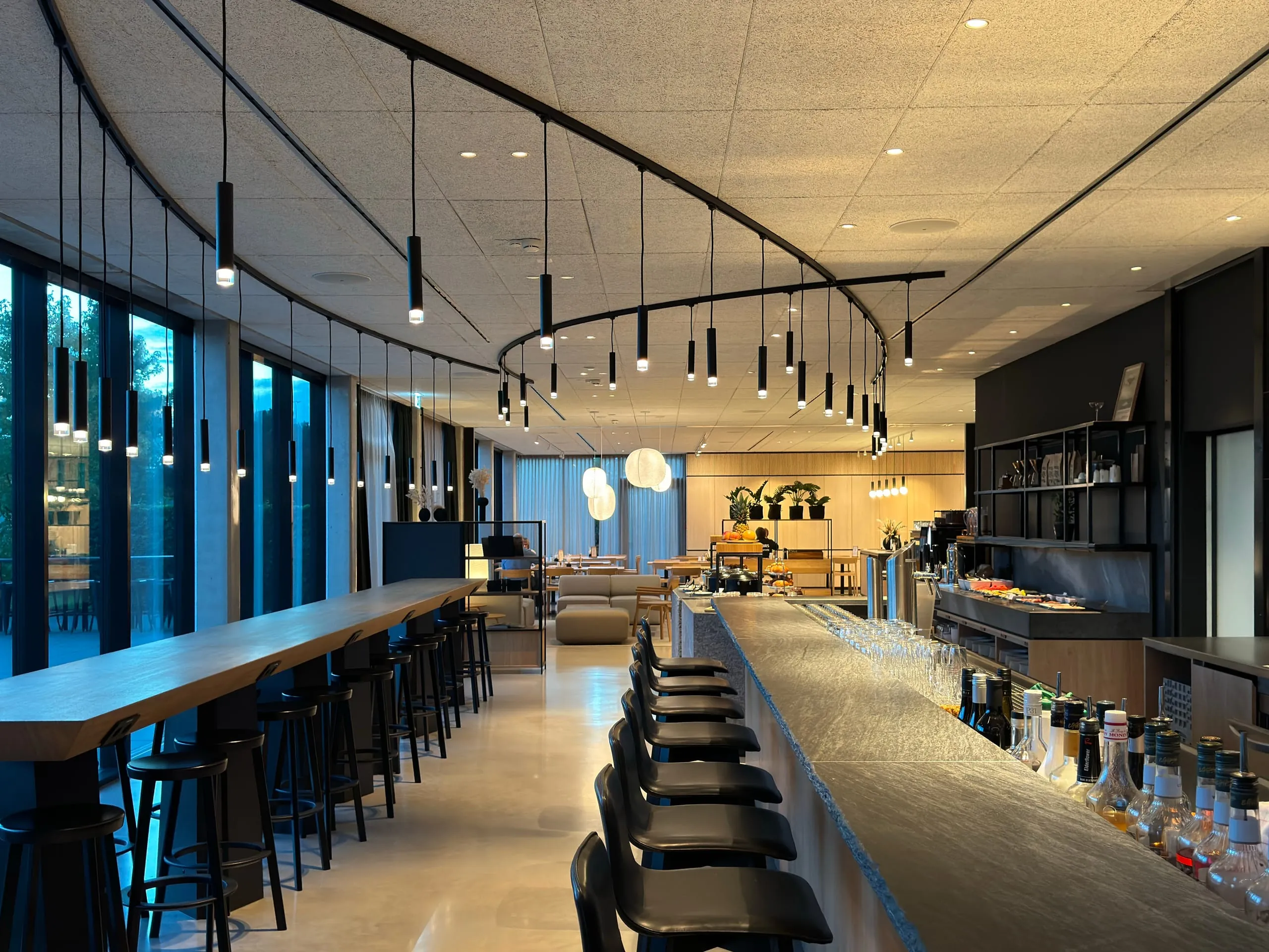 Maßgefertigte Lichtskulptur in der Bar: Pendelleuchten auf gebogenen Schienen setzen dynamische Akzente unter Sichtbeton.