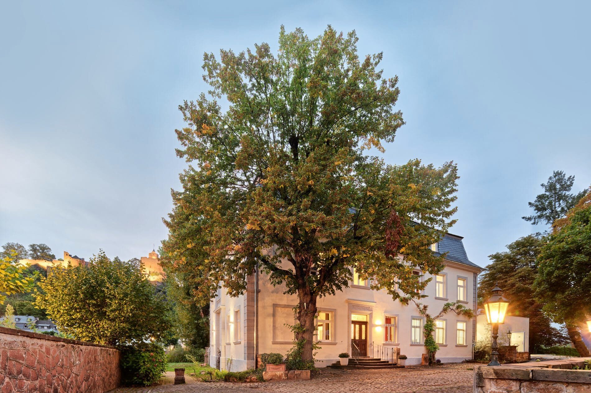 Villa Keller Außenansicht bei Dämmerung mit großem Baum im Innenhof und beleuchteten Fenstern