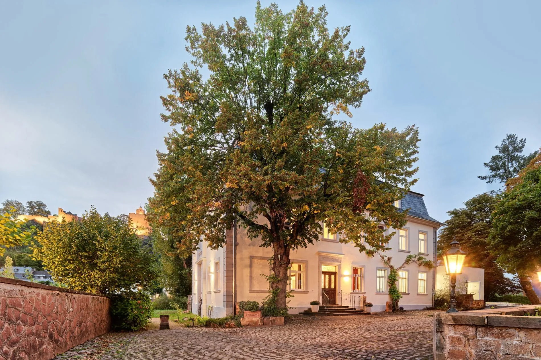 Villa Keller Außenansicht bei Dämmerung mit großem Baum im Innenhof und beleuchteten Fenstern