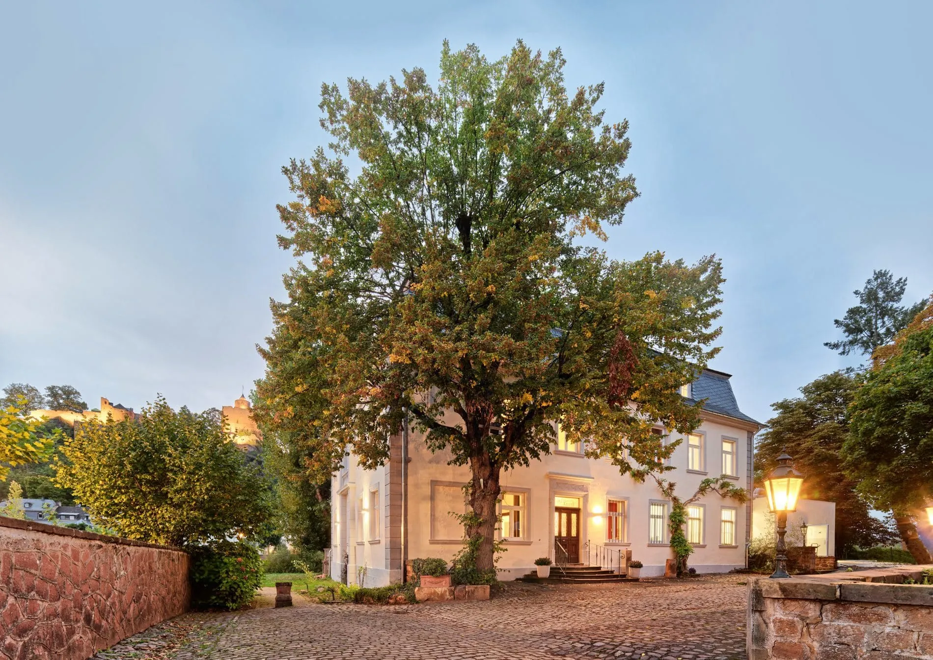 Villa Keller Außenansicht bei Dämmerung mit großem Baum im Innenhof und beleuchteten Fenstern