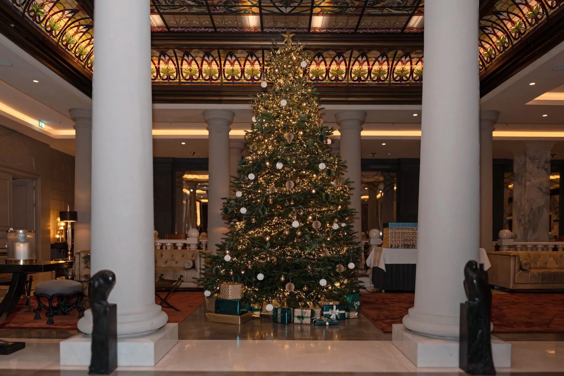 Festlich geschmückter Weihnachtsbaum im Atrium des TITANIC Gendarmenmarkt Berlin mit goldenen Lichtern und Geschenken.