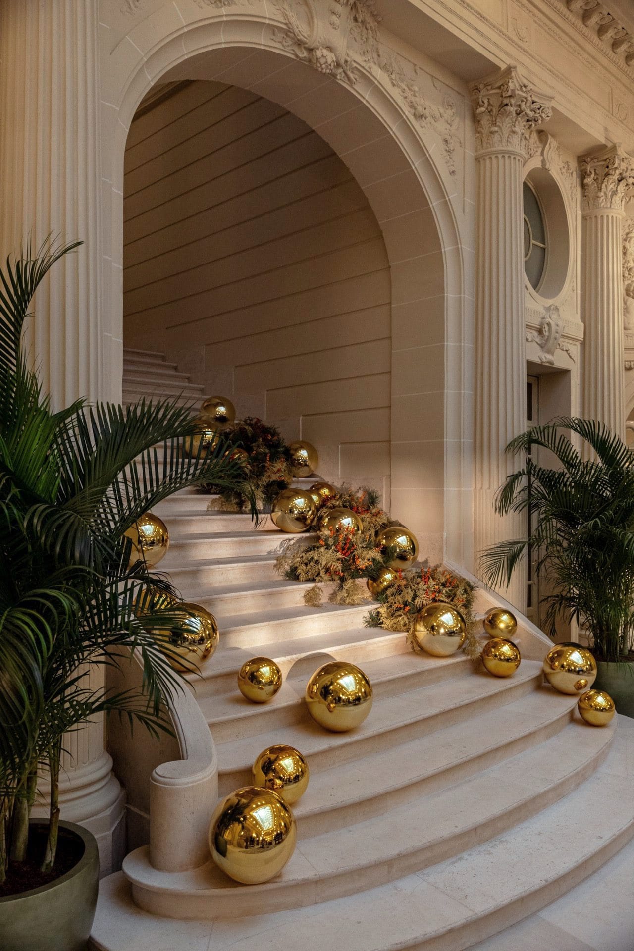 Marmortreppe mit goldenen Weihnachtskugeln, Blumendeko und Palmen in eleganter Hotellobby mit korinthischen Säulen.
