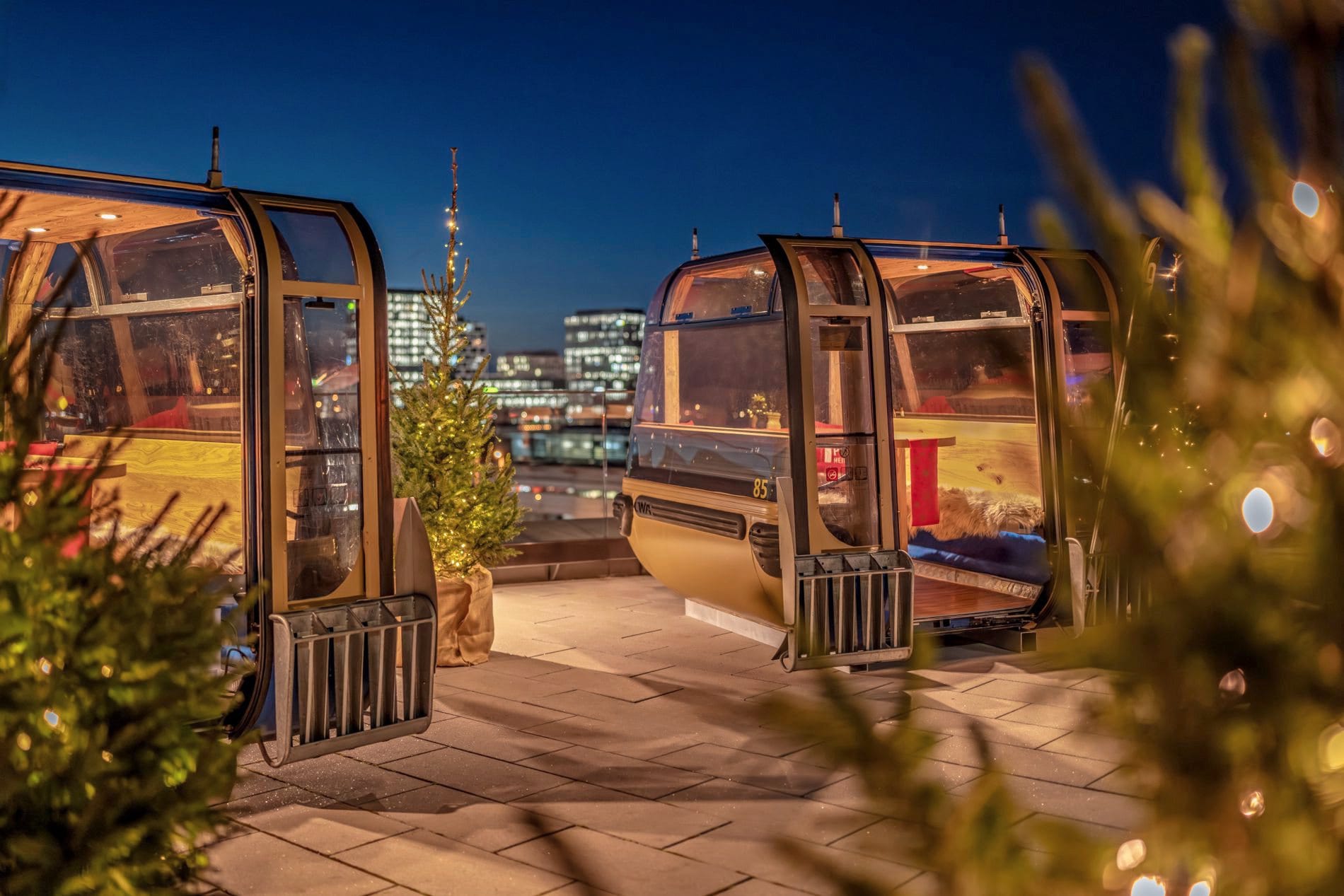Beleuchtete Skigondeln auf Dachterrasse mit Weihnachtsbäumen und Münchner Skyline bei Nacht.
