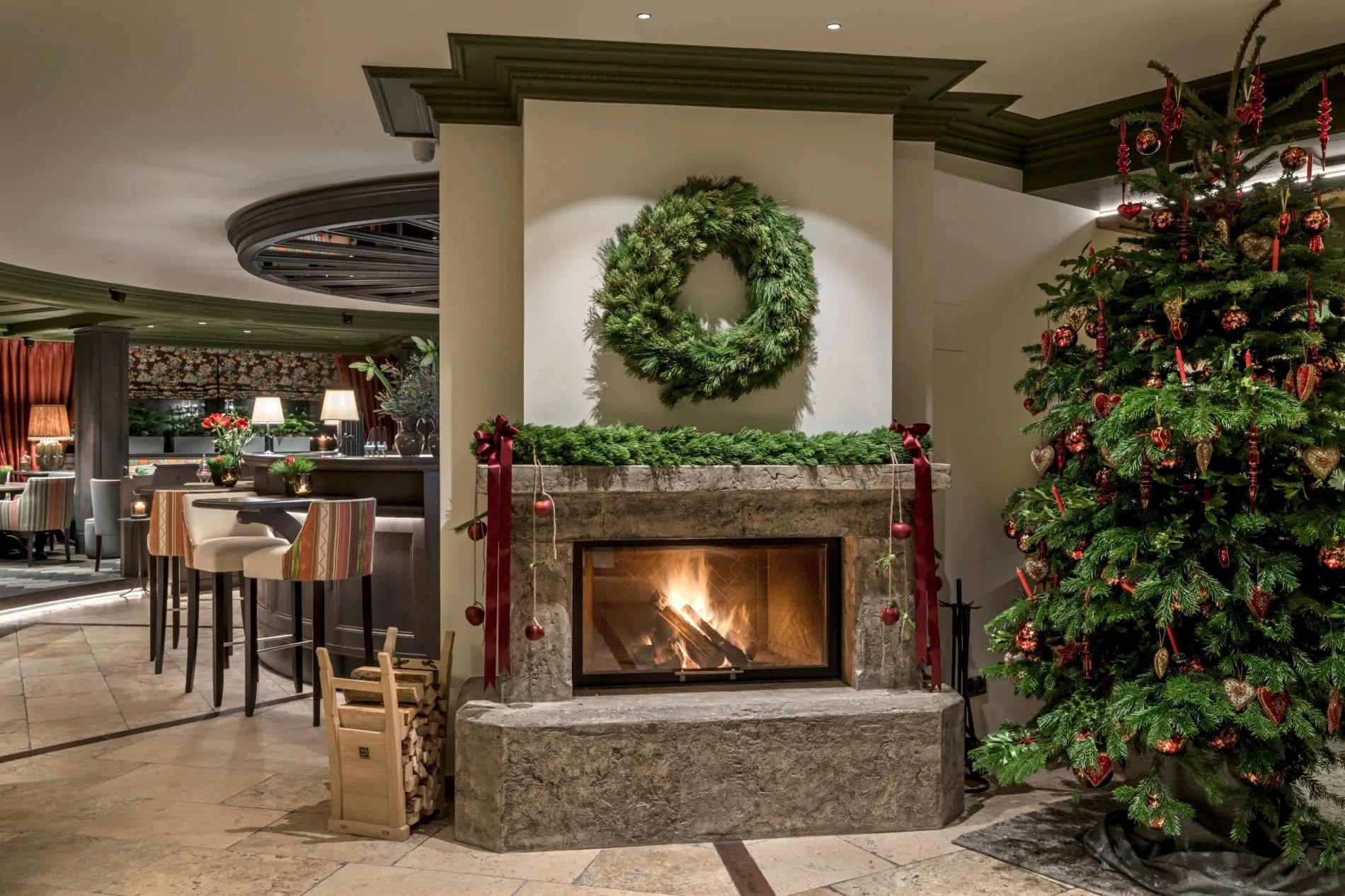 Festliche Atmosphäre in der Hotellobby des Unterschwarzachhofs: Ein lodernder Steinkamin mit Adventskranz, geschmückt mit roten Bändern und Christbaumkugeln, lädt gemeinsam mit dem dekorierten Weihnachtsbaum zum gemütlichen Verweilen in der Vorweihnachtszeit ein.