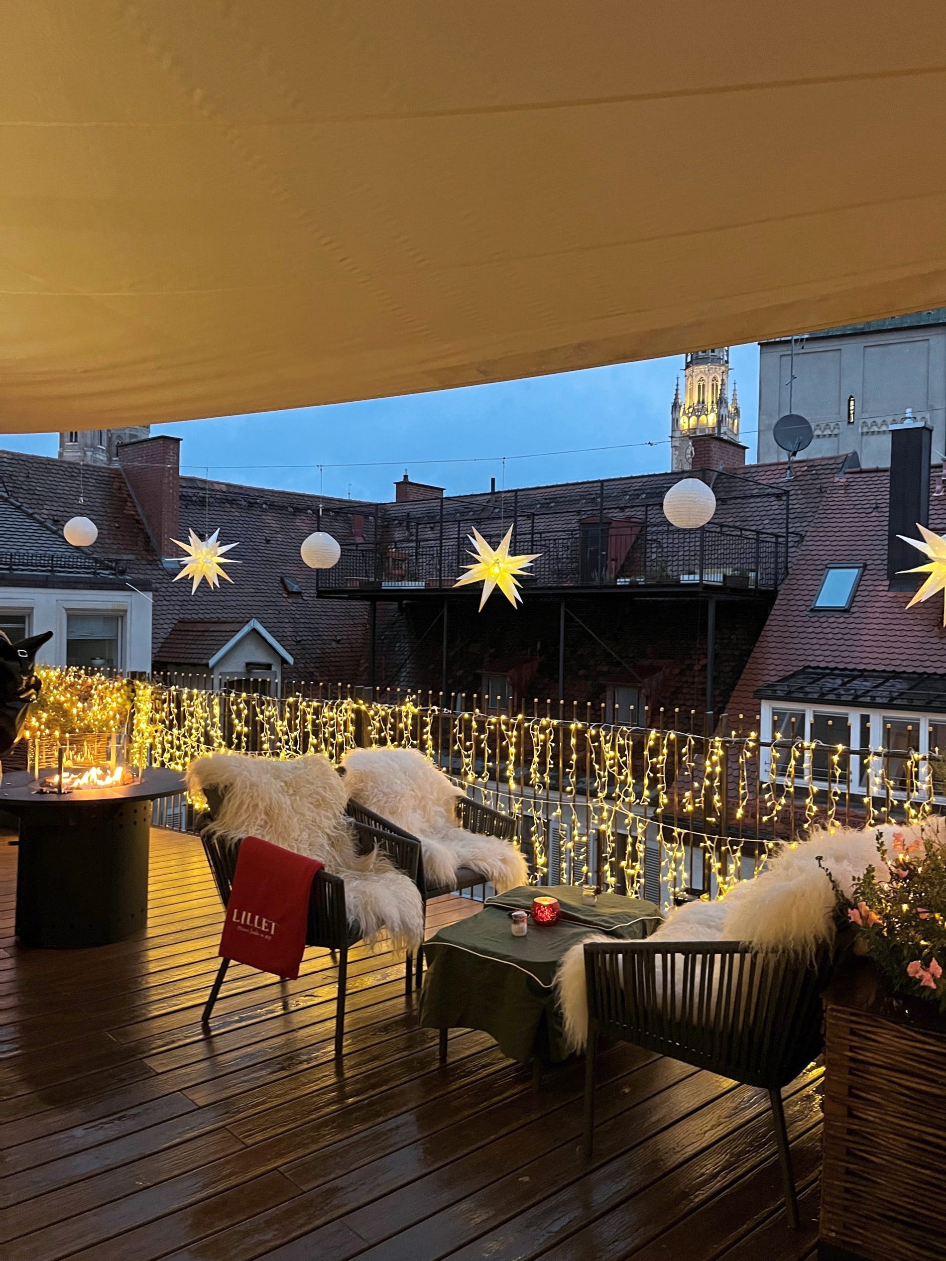 Festlich beleuchtete Dachterrasse mit Lichterketten, Sternen und gemütlichen Sitzplätzen, Blick auf Münchner Altstadt.