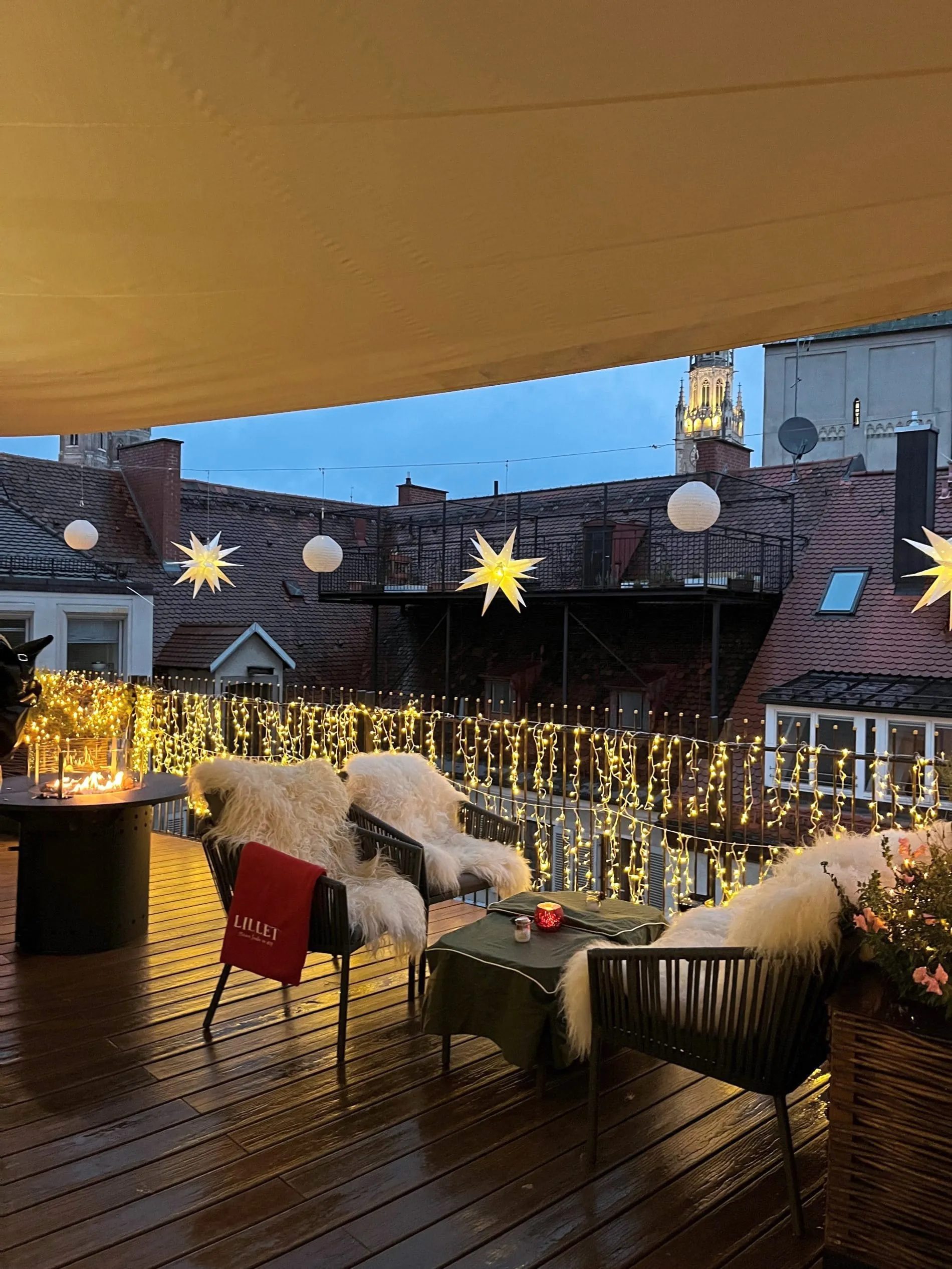 Festlich beleuchtete Dachterrasse mit Lichterketten, Sternen und gemütlichen Sitzplätzen, Blick auf Münchner Altstadt.