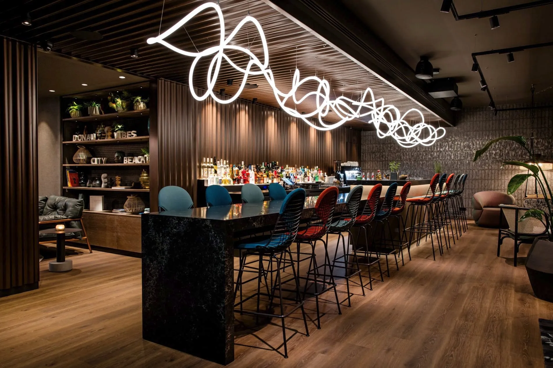 Lange Hotelbar mit bunten Barhockern, weißer Neon-Lichtkunst und Regalen mit Wien-Bezug.