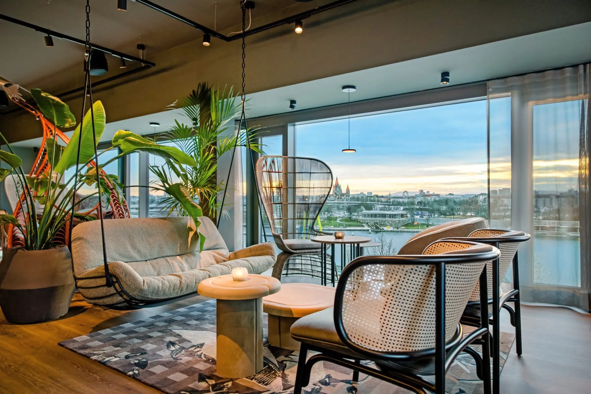 Lounge mit Panoramafenstern zur Donau, Rattanmöbeln, Hängeschaukel und Grünpflanzen.