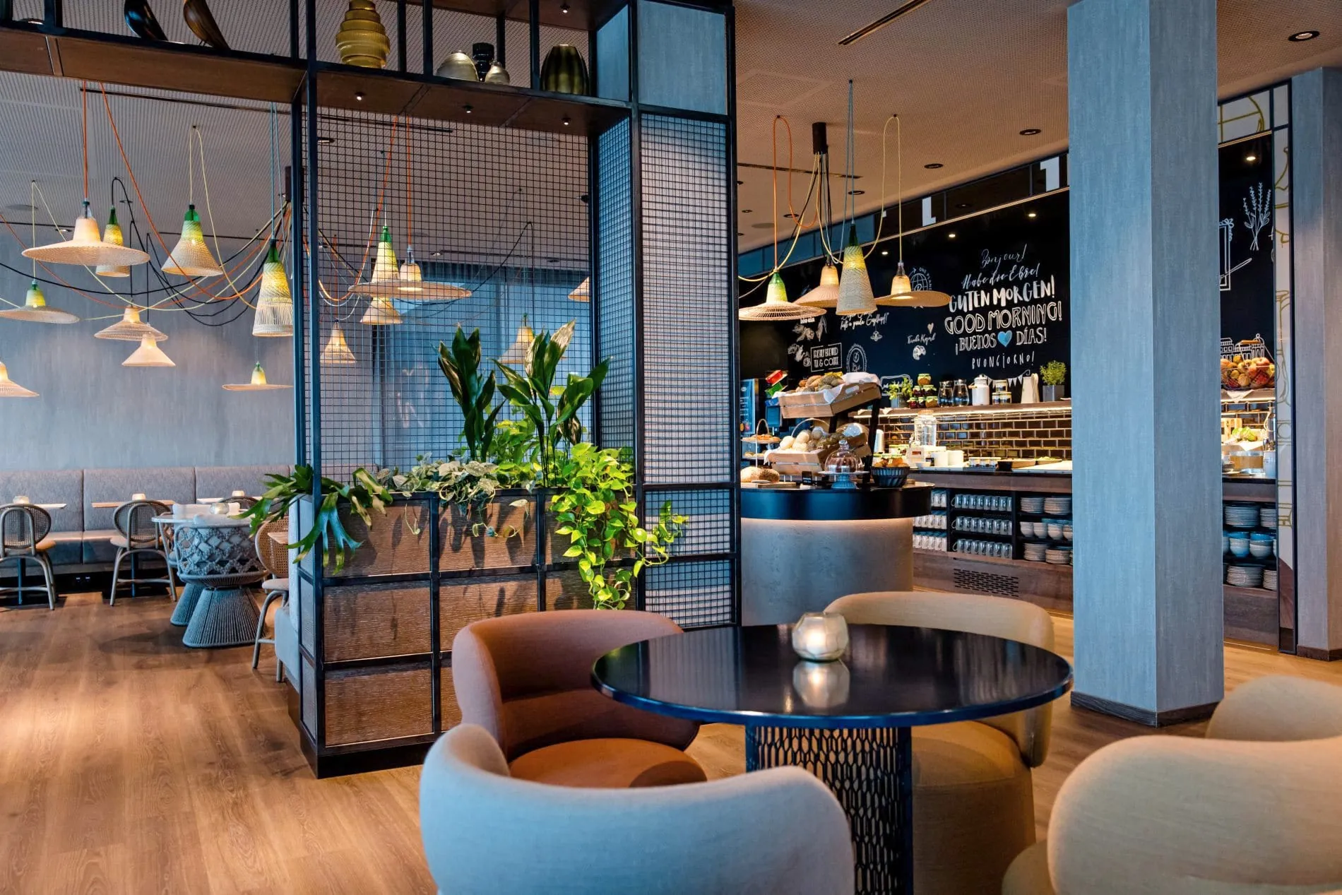 Frühstücksbereich-Bereich mit Glaswänden, Pendelleuchten, bunten Stühlen und Tafelwand mit Begrüßungen.