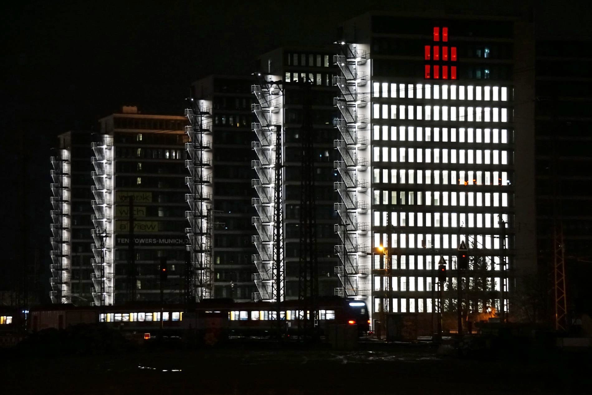 Nächtliche Ansicht der Ten Towers München mit weißer Fassadenbeleuchtung und roter Adventskerzen-Illumination.