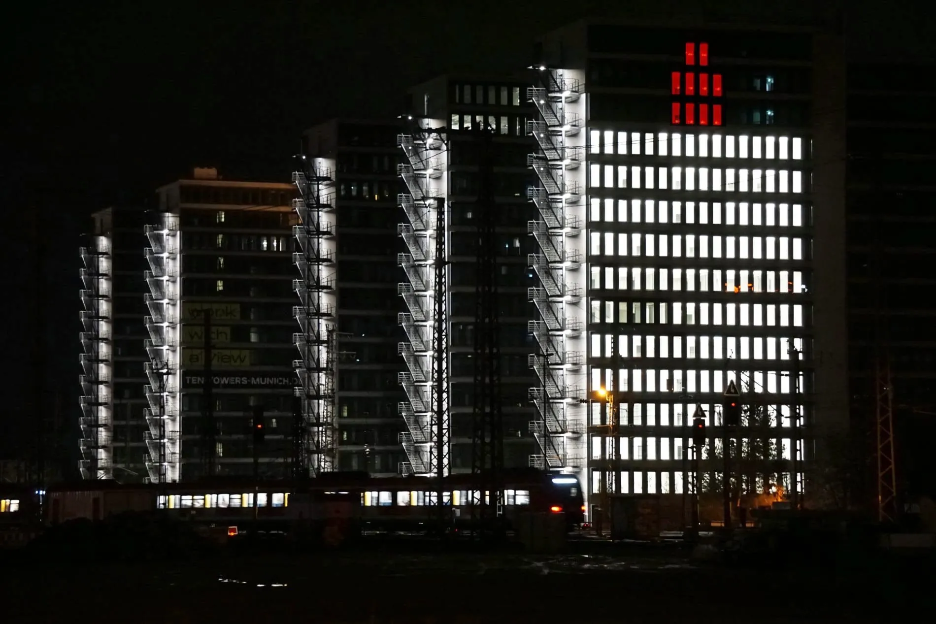 Nächtliche Ansicht der Ten Towers München mit weißer Fassadenbeleuchtung und roter Adventskerzen-Illumination.