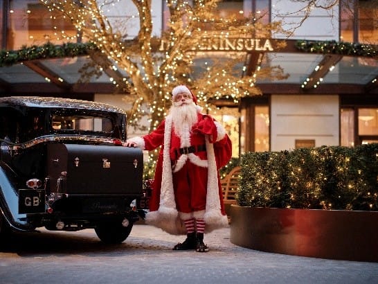 Weihnachtsmann vor dem festlich beleuchteten Eingang von The Peninsula London mit Oldtimer und Lichterdekoration.