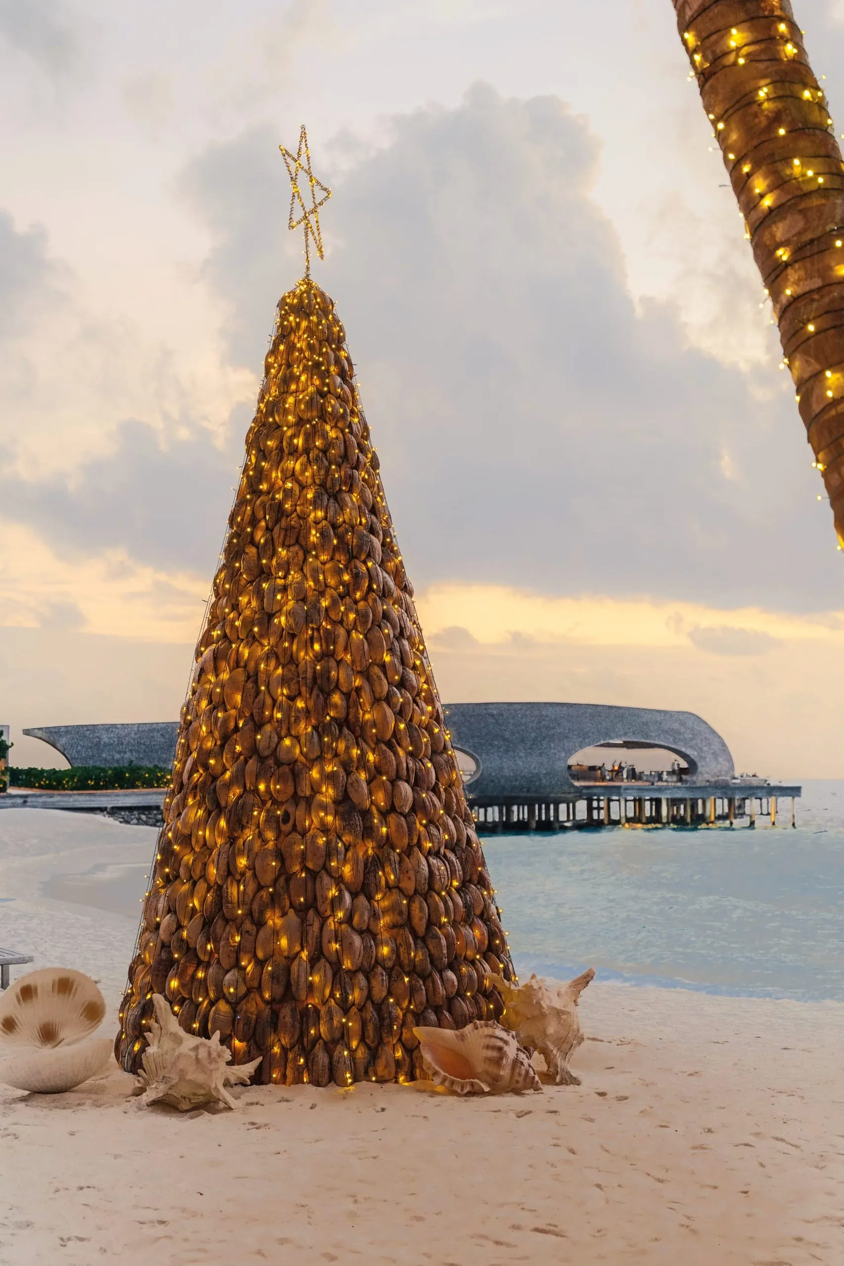 Beleuchteter Weihnachtsbaum aus Kokosnüssen am Strand mit Luxusresort-Villen über dem Wasser im Hintergrund.