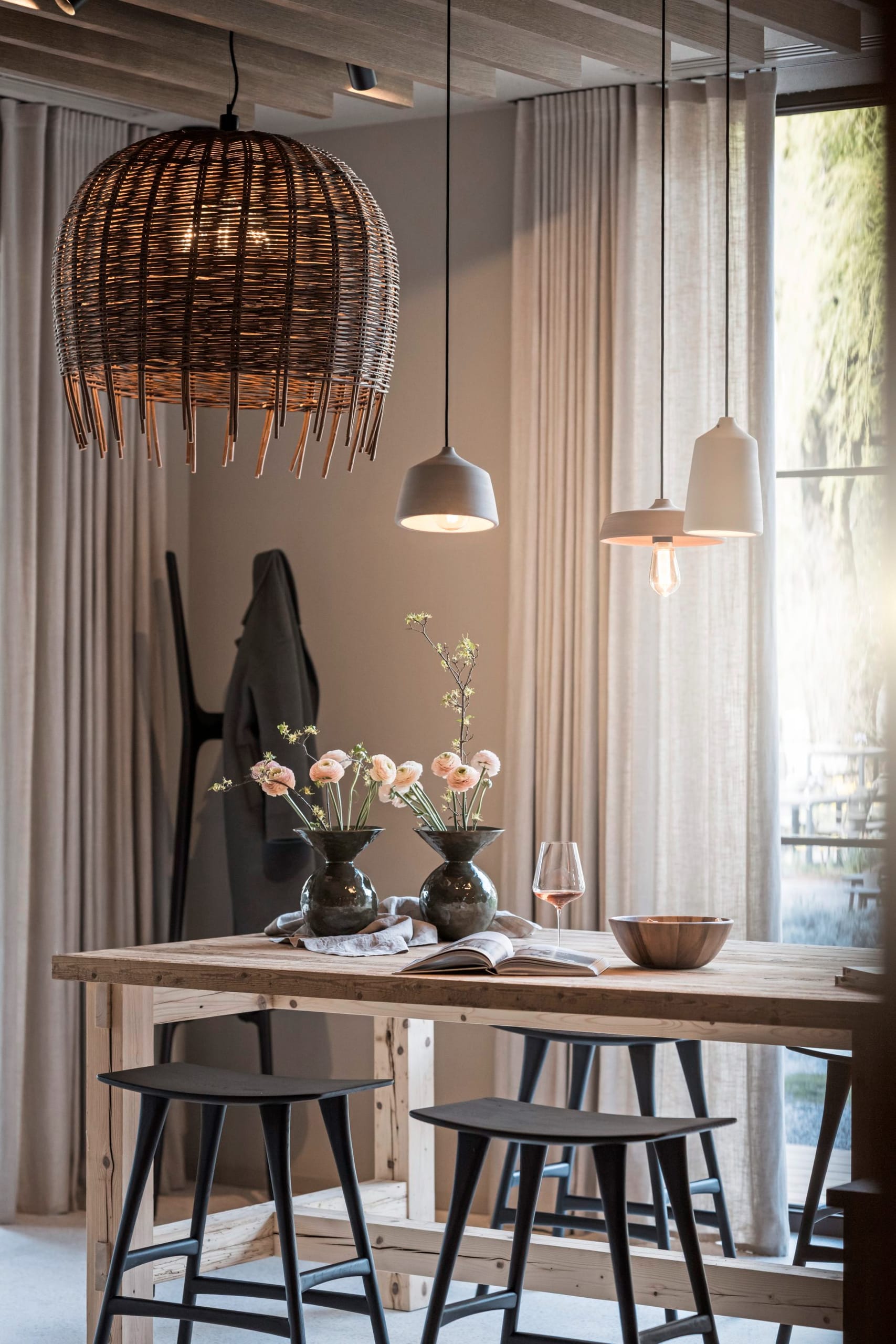 Holztisch mit Blumenvasen, Weinglas und Rattanleuchte, schwarze Hocker.