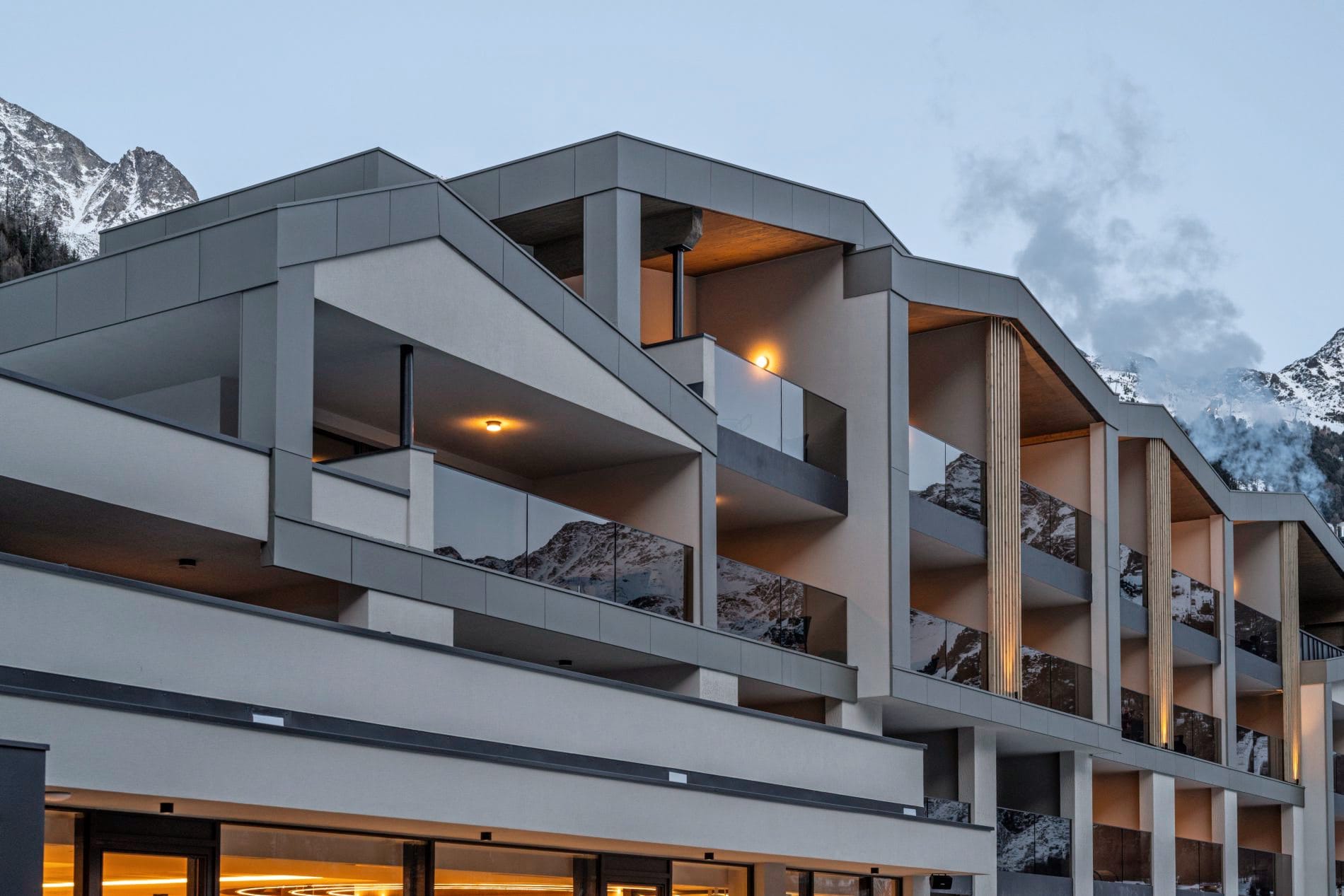 Moderne Hotelfassade mit mehreren Balkonen, Glasgeländern und Bergpanorama im Hintergrund.