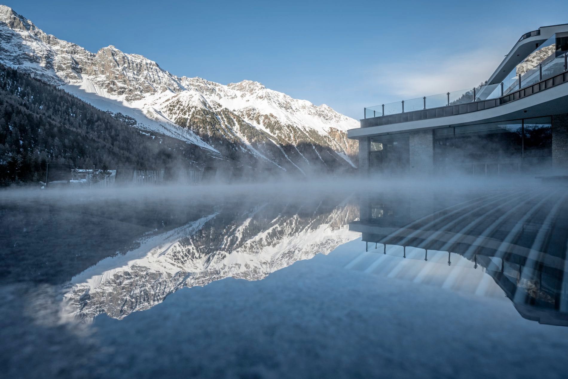 Beheizter Außenpool mit aufsteigendem Dampf und Spiegelung der schneebedeckten Berge