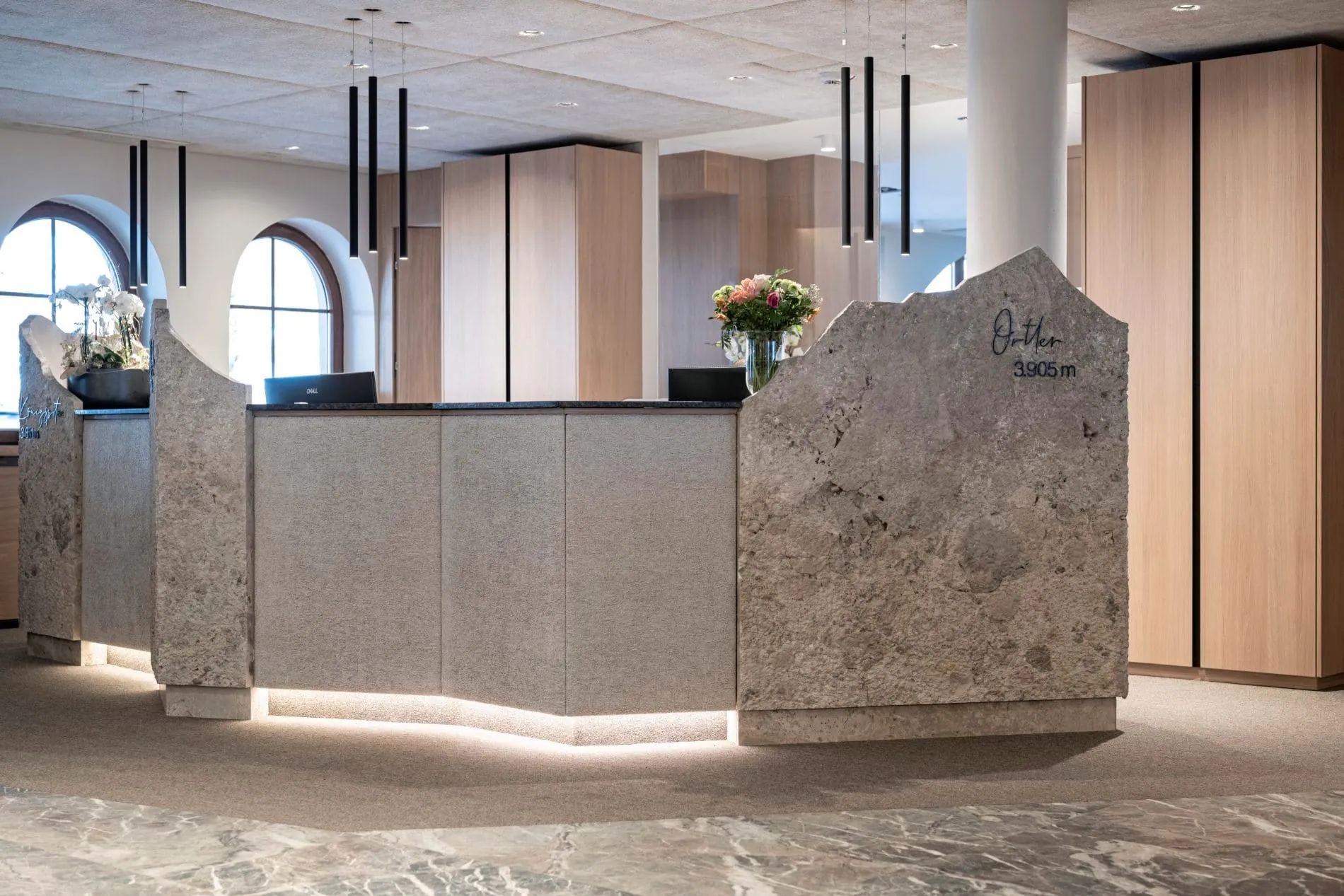 Hotelrezeption mit grauem Natursteintresen in Bergform und Holzelementen.