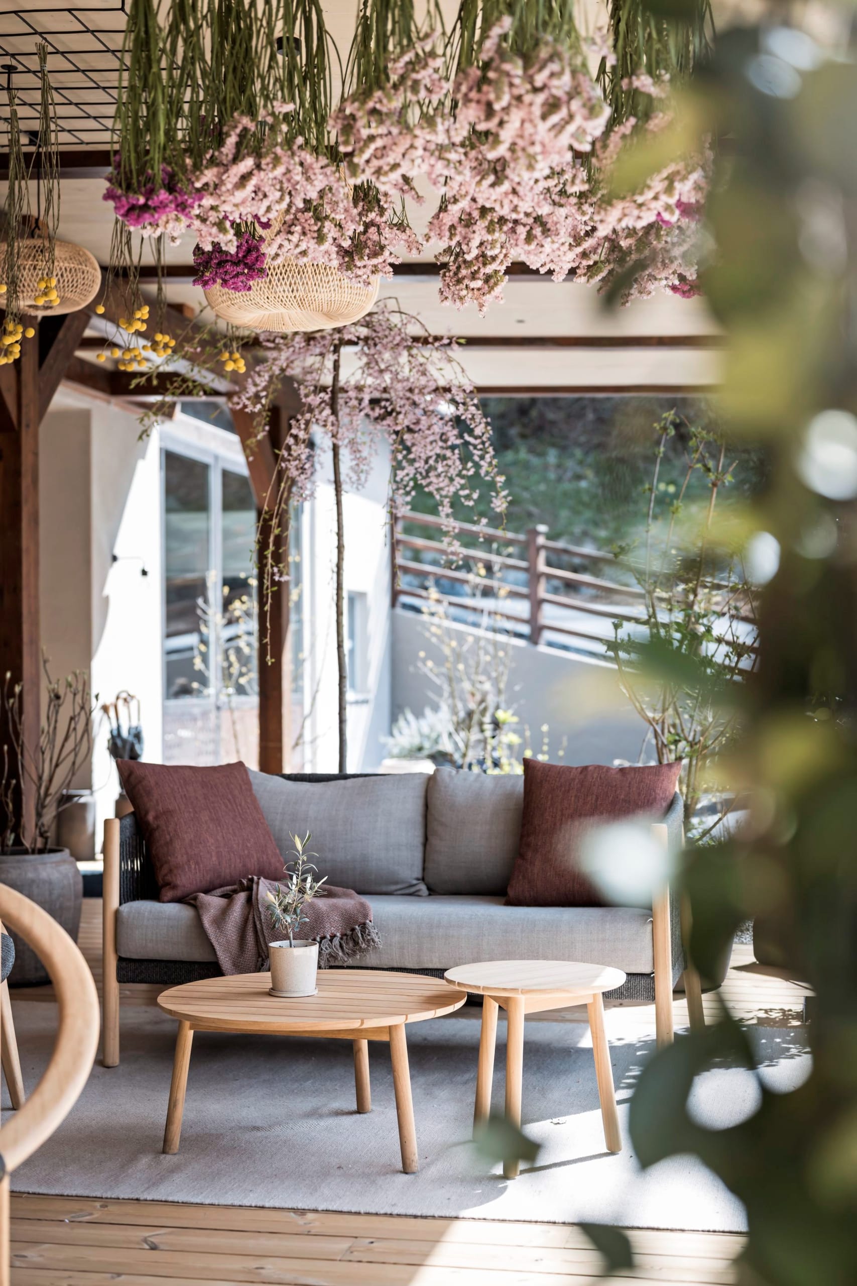Outdoor-Sofa mit Kissen unter hängenden rosa Blüten und Holztischen.