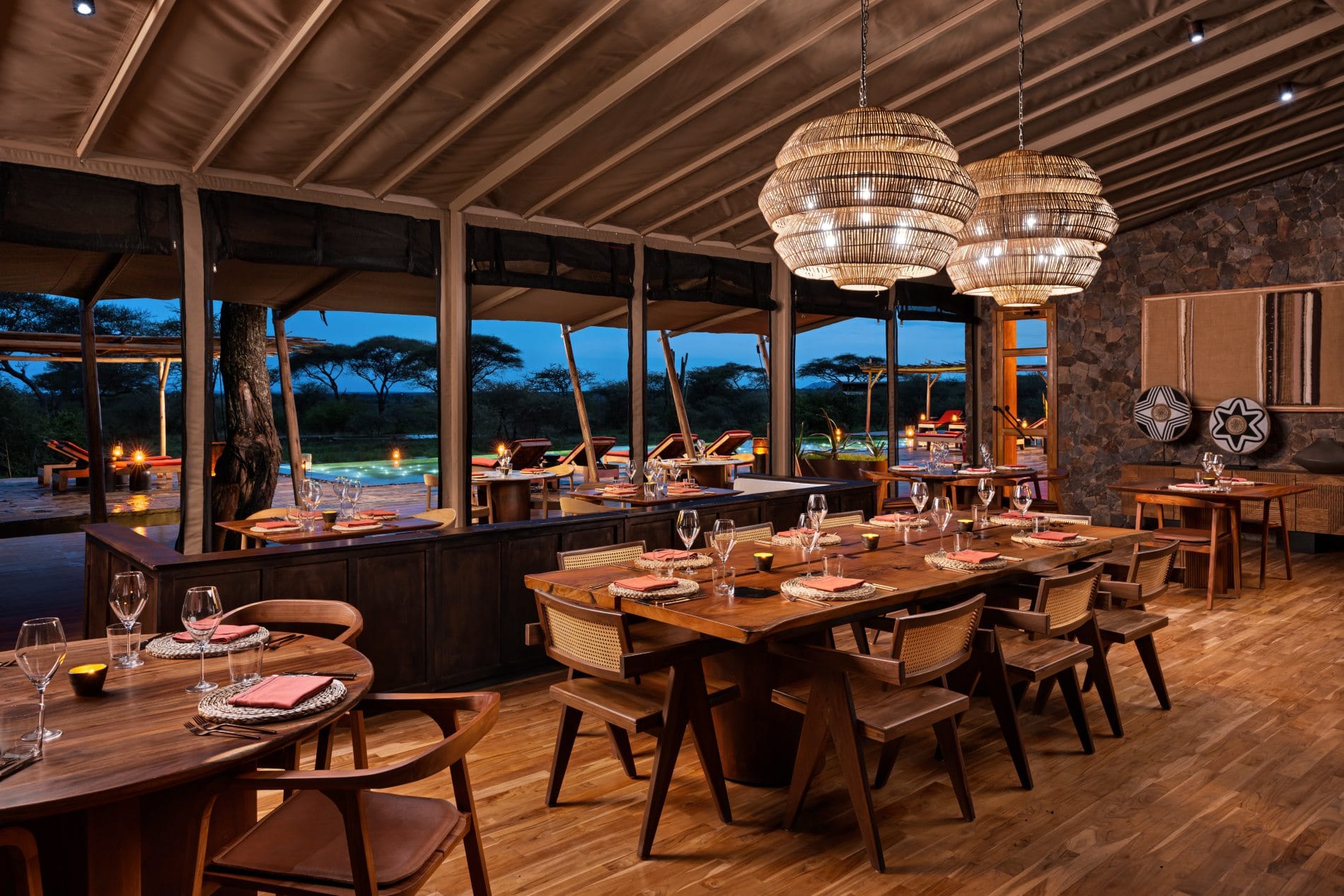 Elegant gedeckte Holztische im Restaurant mit Rattanlampen und Blick auf den beleuchteten Pool.