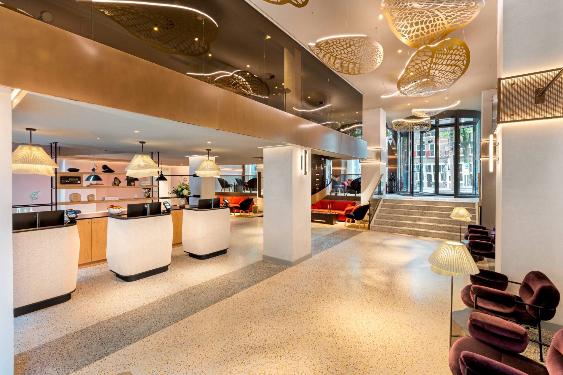 Moderne Hotellobby mit Empfangstresen, goldenen Deckenskulpturen, Terrazzoboden und Glaseingang.