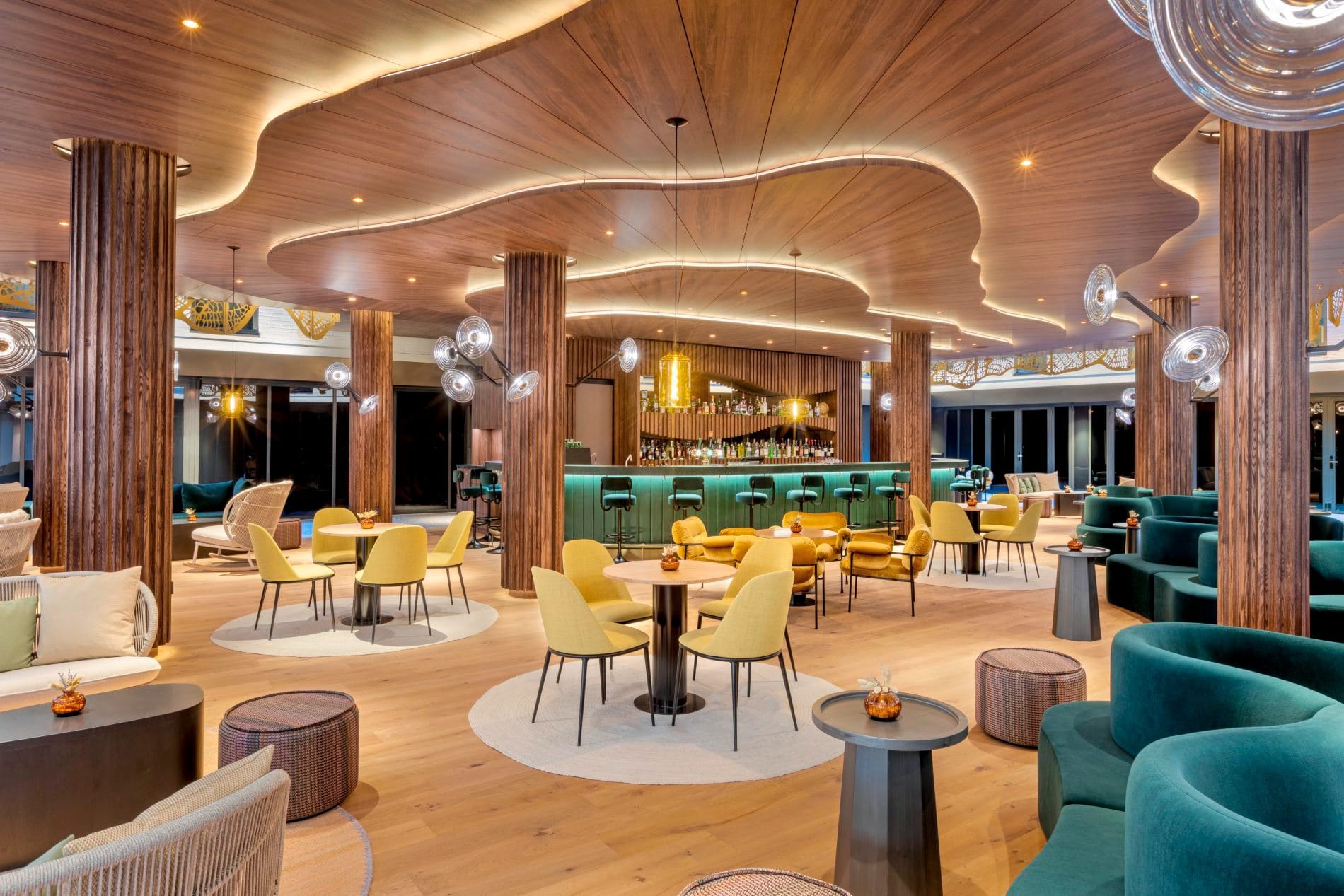 Elegante Hotelbar mit geschwungener Holzdecke, grüner Theke und bunten Sesseln in Gelb und Petrol.