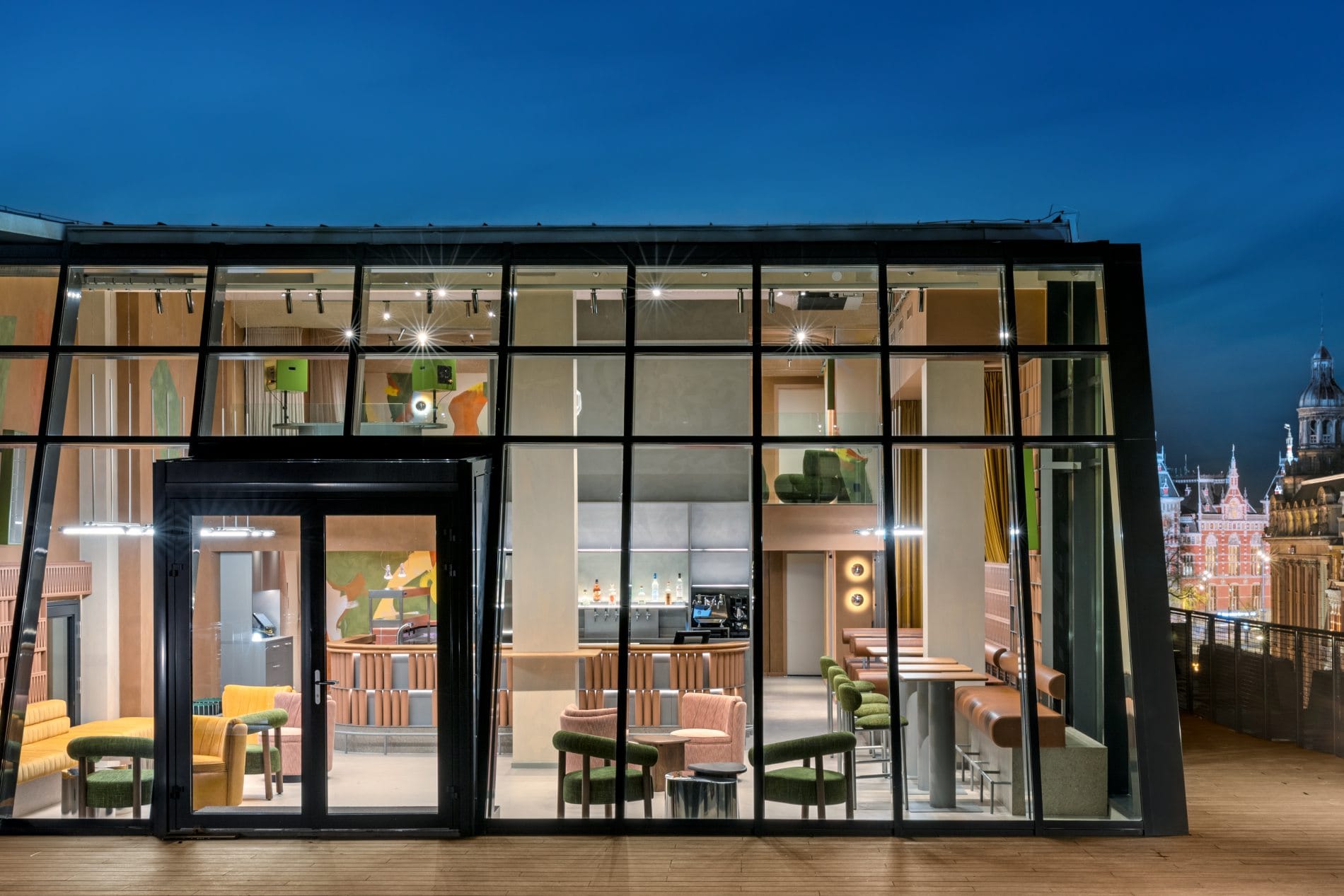 Verglaste Rooftop-Bar bei Nacht mit Blick auf historische Amsterdamer Gebäude und Holzterrasse.