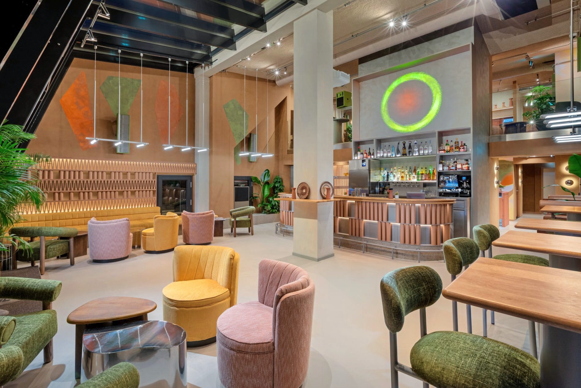 Hotelbar mit bunten Sesseln in Gelb, Rosa und Grün, Bar mit Getränkeregal und Neon-Kunstwerk.