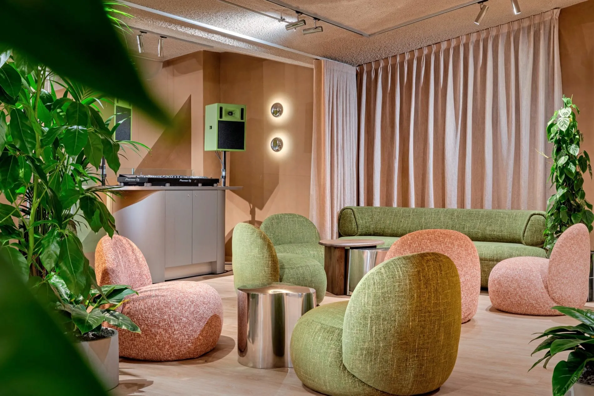 Moderne Hotellounge mit DJ-Pult, organischen Sesseln in Grün und Rosa, Grünpflanzen und Vorhängen.