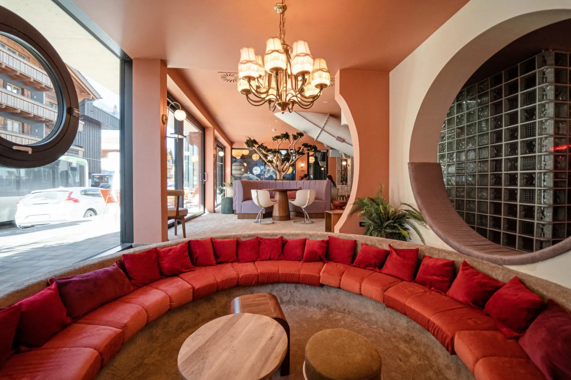 Hotel-Lounge mit rotem Rundsofa, Kronleuchter, rundem Fenster und Blick auf Berglandschaft.