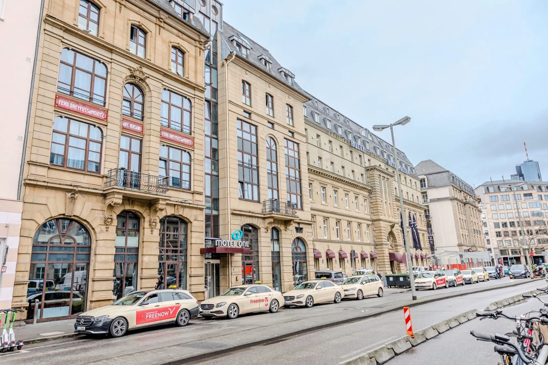 Außenansicht des Motel One Frankfurt-Hauptbahnhof in historischer Sandsteinfassade mit Taxis davor.