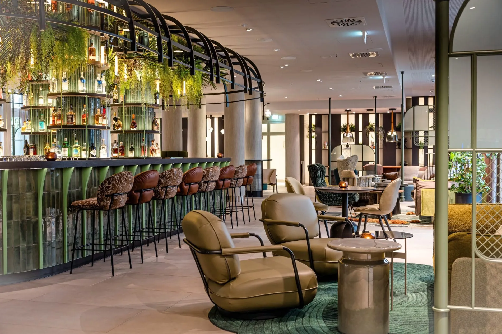 Bar im Motel One Frankfurt mit grünen Glasregalen, Spirituosen, Lederbarhockern und Pflanzen.