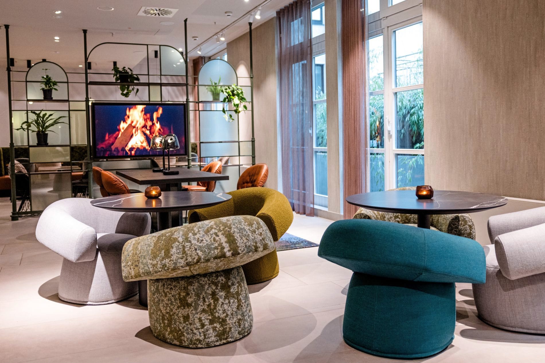 Lounge im Motel One Frankfurt mit bunten Polstersesseln, Beistelltischen und Kaminbildschirm.