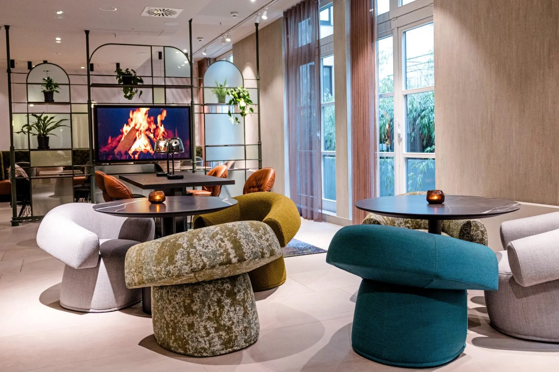 Lounge im Motel One Frankfurt mit bunten Polstersesseln, Beistelltischen und Kaminbildschirm.