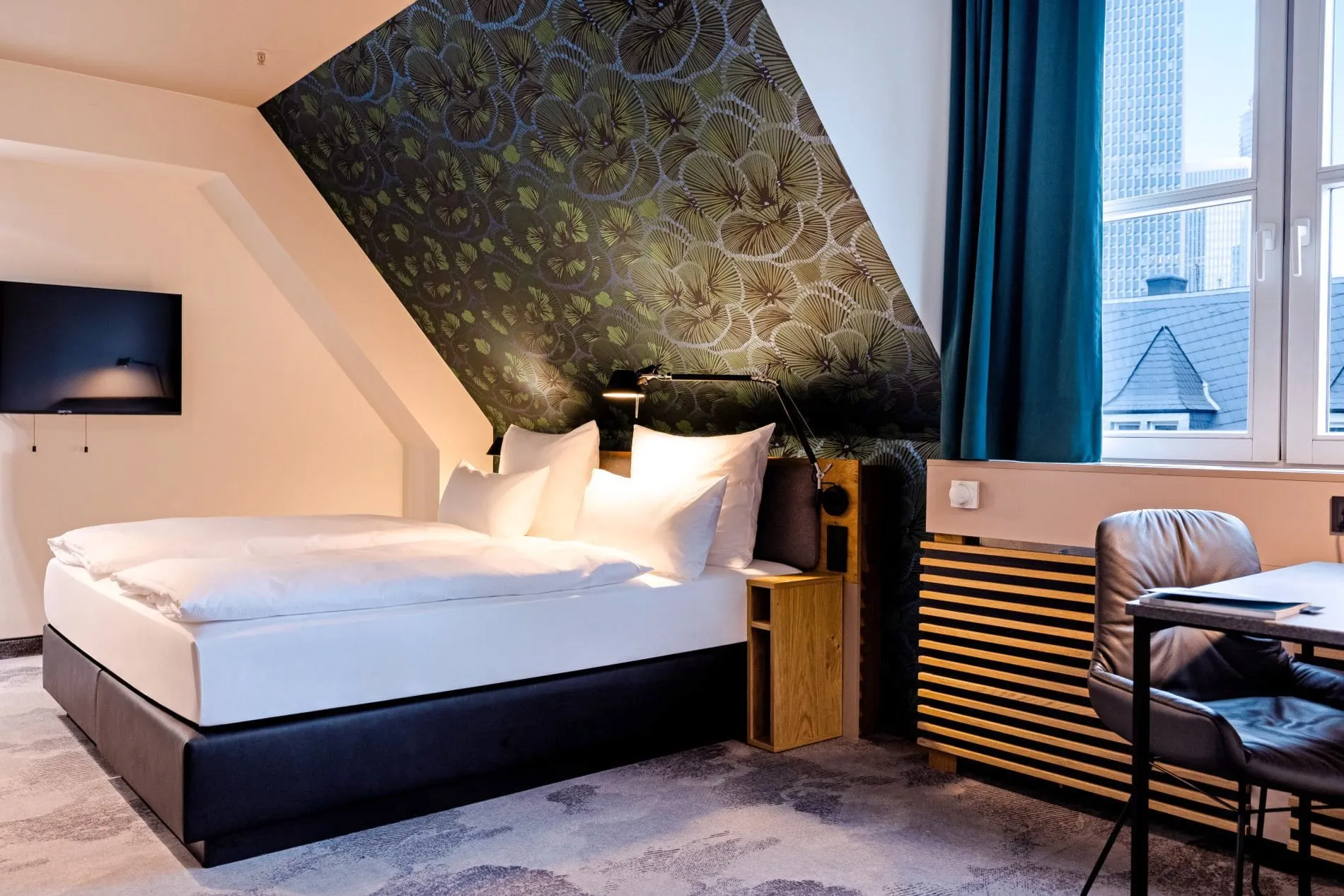 Hotelzimmer im Motel One Frankfurt mit Doppelbett, Dachschräge, gemusterter Tapete und Schreibtisch.