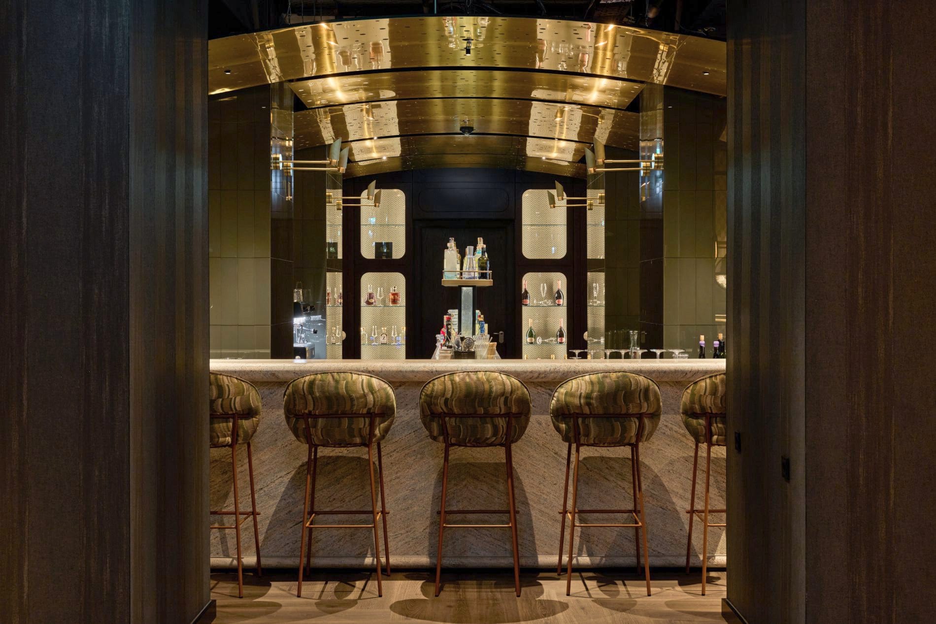 Hotelbar mit vier Barhockern, Marmortresen, goldener Deckenverkleidung und beleuchteten Flaschenregalen.