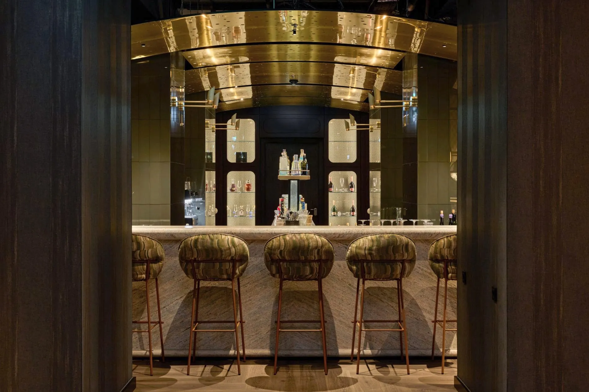 Hotelbar mit vier Barhockern, Marmortresen, goldener Deckenverkleidung und beleuchteten Flaschenregalen.