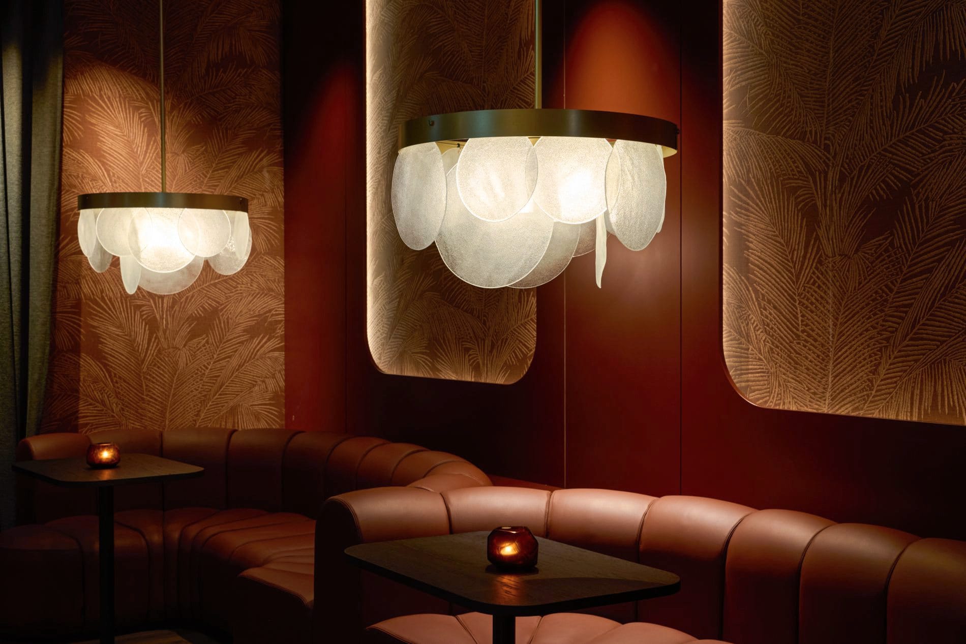 Lounge mit roten Ledersofas, Palmenmuster-Tapete und blütenförmigen Pendelleuchten im warmen Licht.