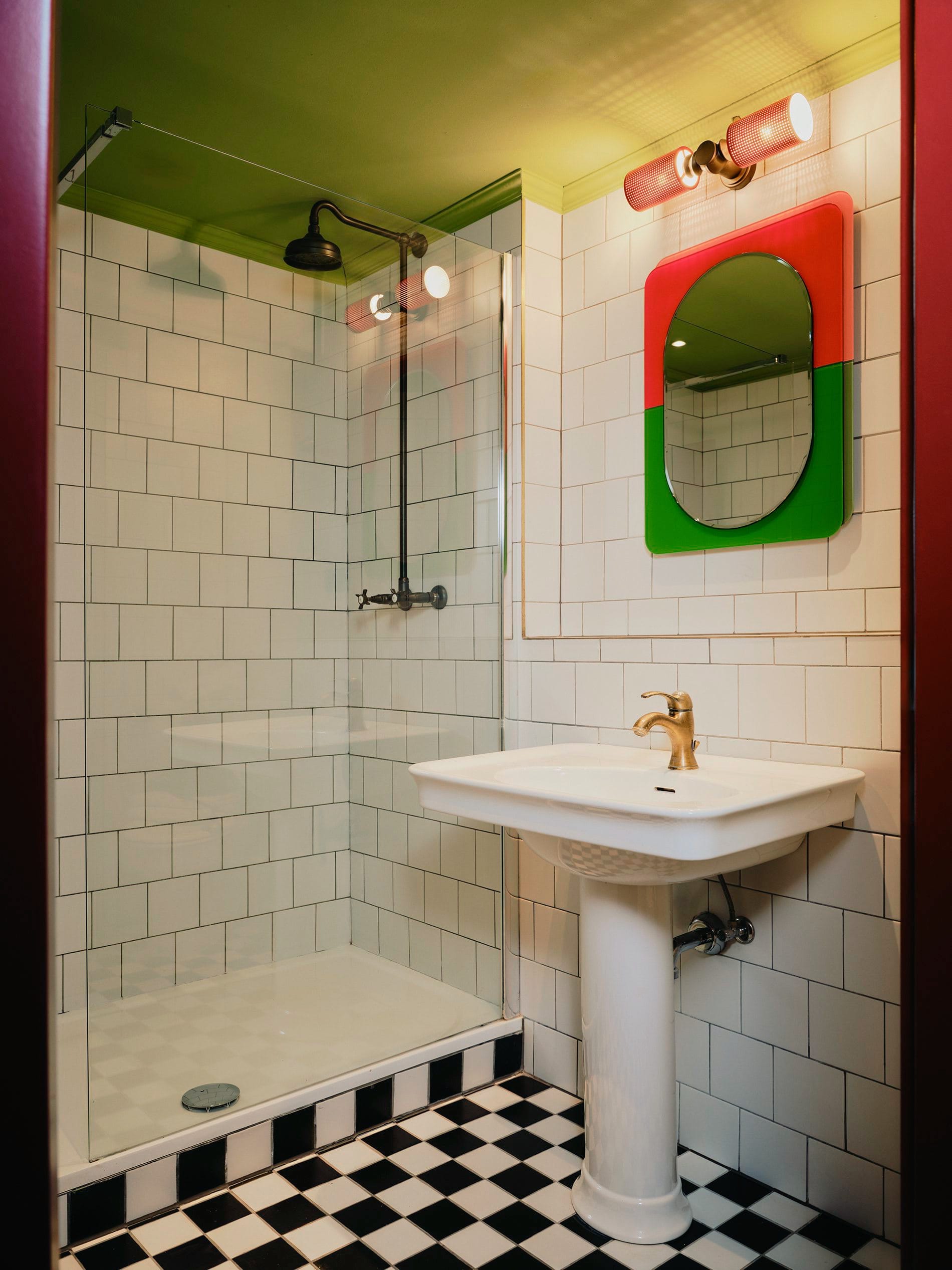 Historische Fliesenarbeiten treffen auf kräftige Farbakzente. Der rot-grüne Spiegel und die olivgrüne Decke setzen spielerische Kontraste zu den klassischen weißen Metrofliesen und dem Schachbrettboden.