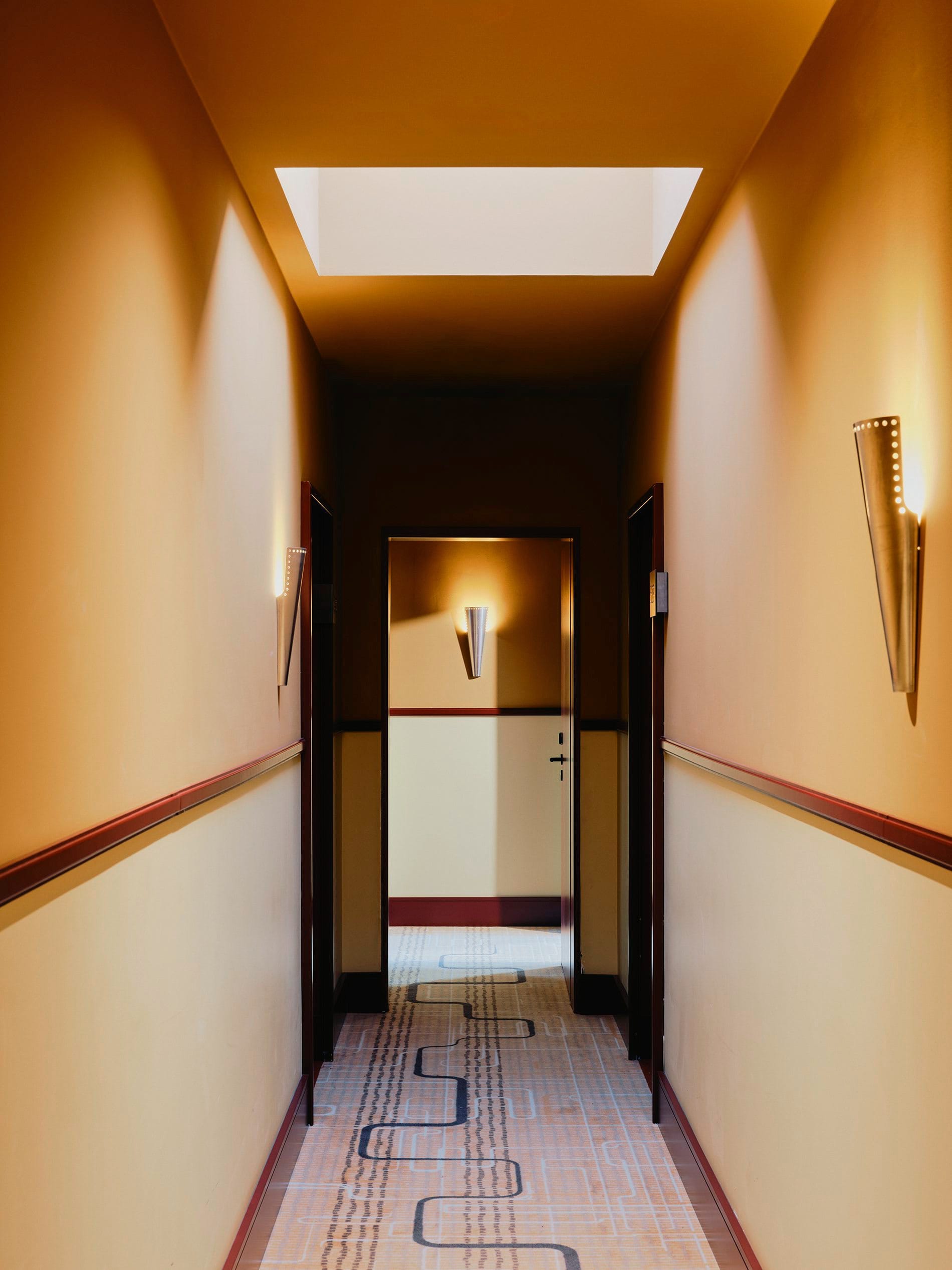 Skulpturale Wandleuchten in Kegelform und ein gemusterter Teppich mit geschwungenen Linien führen durch die Flure des historischen Gebäudes. Die warmen Ockertöne schaffen eine einladende Atmosphäre.