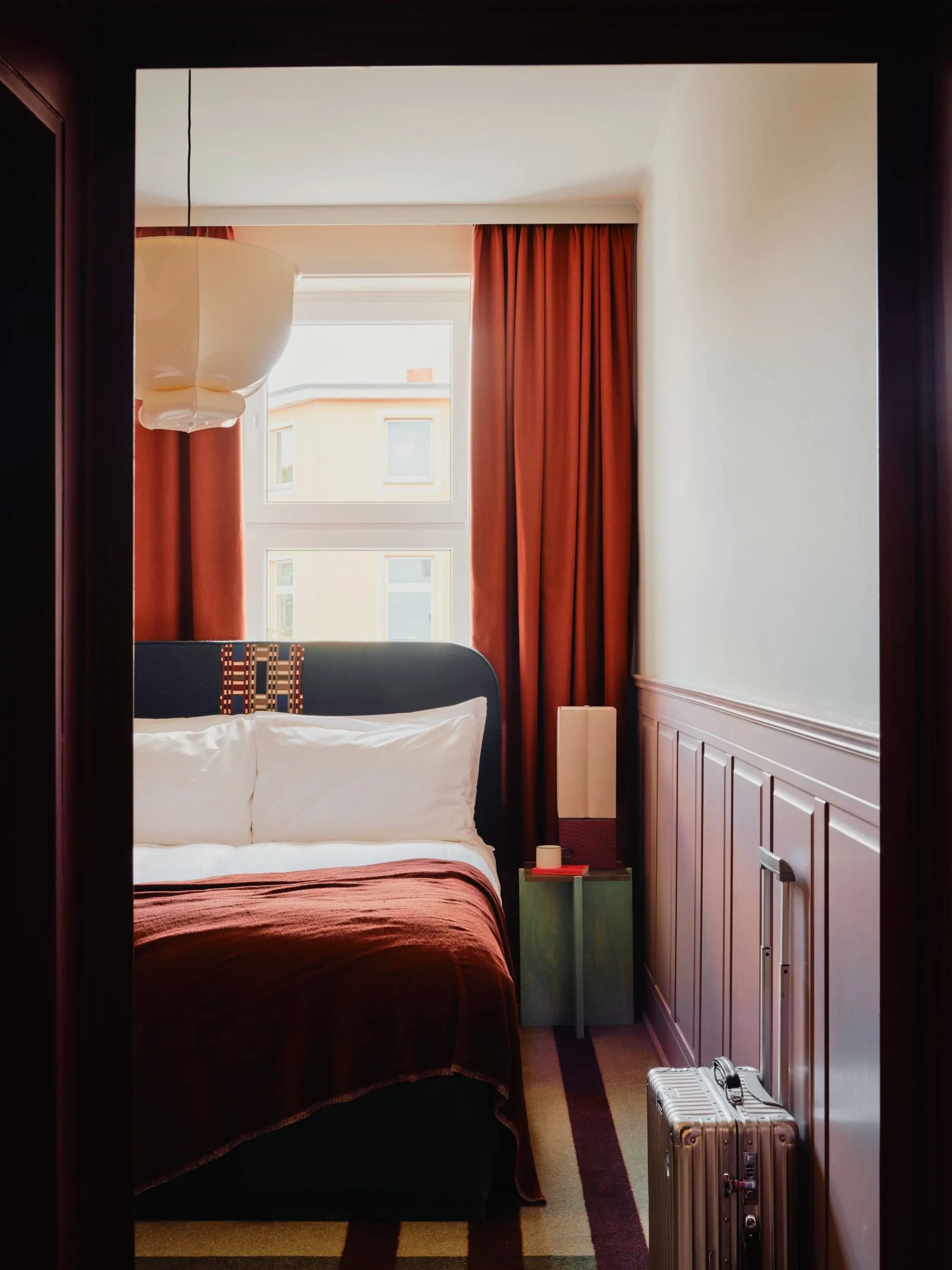 Der tiefrot gestrichene Eingangsbereich rahmt den Blick auf das Doppelbett mit mauvefarbener Holzvertäfelung. Geschichtete Textilien und raumhohe Vorhänge prägen das warme Farbkonzept der Zimmer.