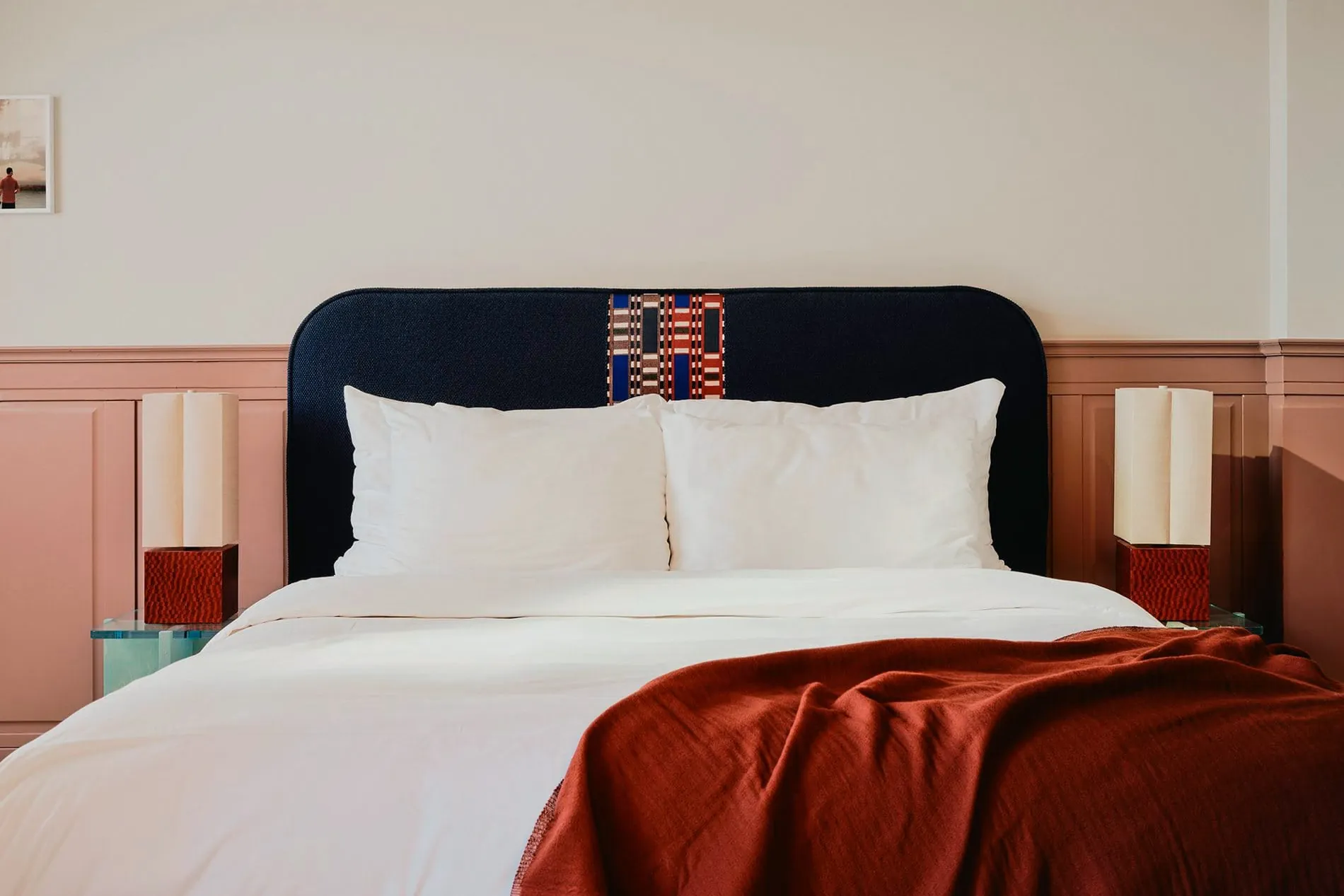 Maßgefertigte Möbel und geschichtete Textilien prägen die Zimmergestaltung. Das dunkelblaue Kopfteil mit eingewebtem Muster wird von symmetrisch platzierten Nachttischleuchten und einer rostfarbenen Tagesdecke ergänzt.