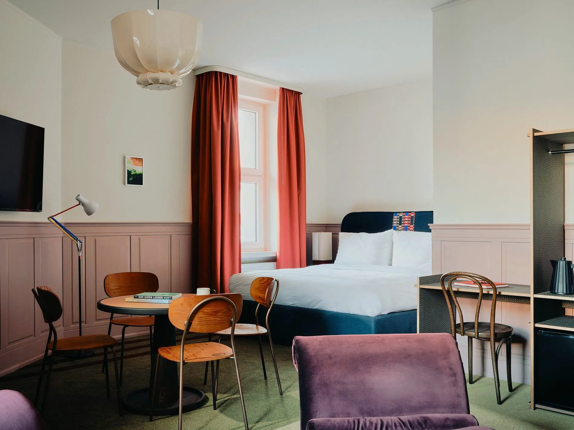 Die größeren Zimmerkategorien bieten separate Sitzbereiche mit rundem Tisch und Vintage-Stühlen. Mauvefarbene Wandvertäfelungen und ein grüner Teppich setzen das charakteristische Farbkonzept fort.