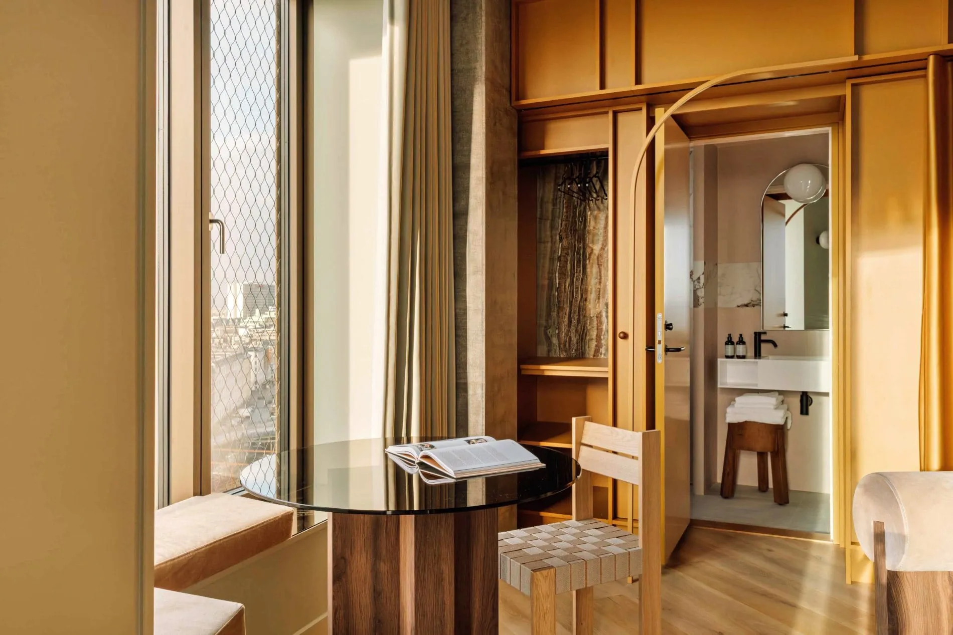 Ein offener Garderobenschrank und der Durchgang zum Badezimmer sind in eine senfgelbe Wandverkleidung integriert. Der runde Glastisch am Fenster dient als Arbeits- und Essplatz mit Blick über die Dächer Kopenhagens.