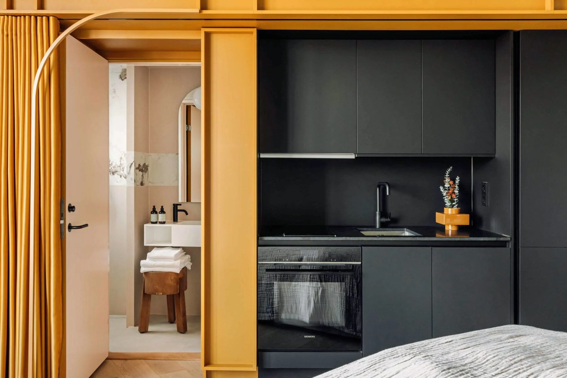 Die Apartments verfügen über eine kompakte Kitchenette in mattem Schwarz und einen senfgelben Vorhang, der den Übergang zum marmorgefliesten Badezimmer markiert.
