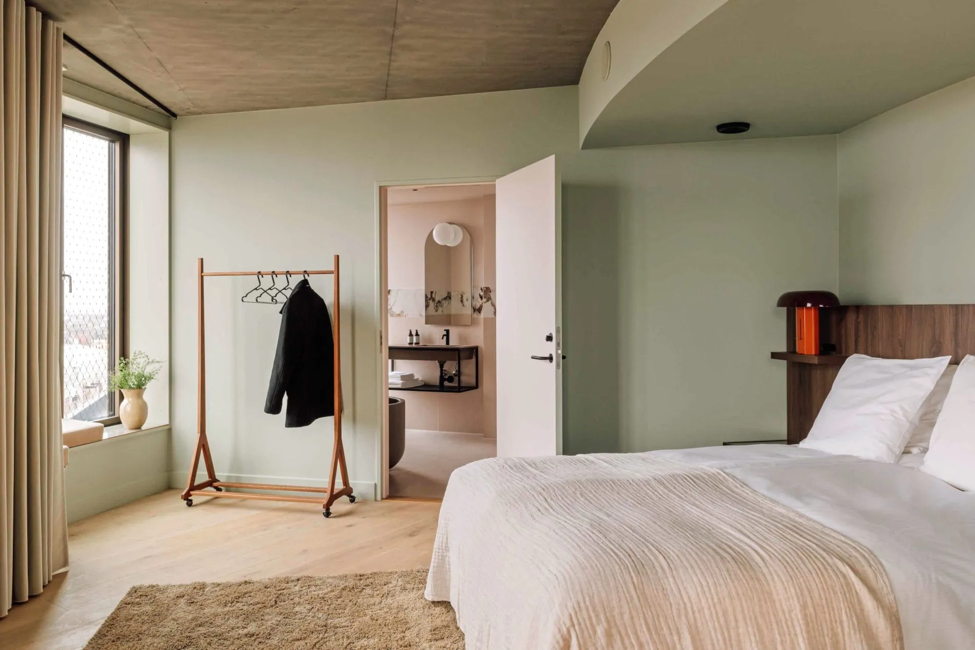 Die Sichtbetondecke verleiht dem Schlafbereich der Suite eine besondere Raumwirkung. Ein offener Kleiderständer aus Holz und der Durchgang zum Badezimmer unterstreichen das Konzept aus Offenheit und Wohnlichkeit.