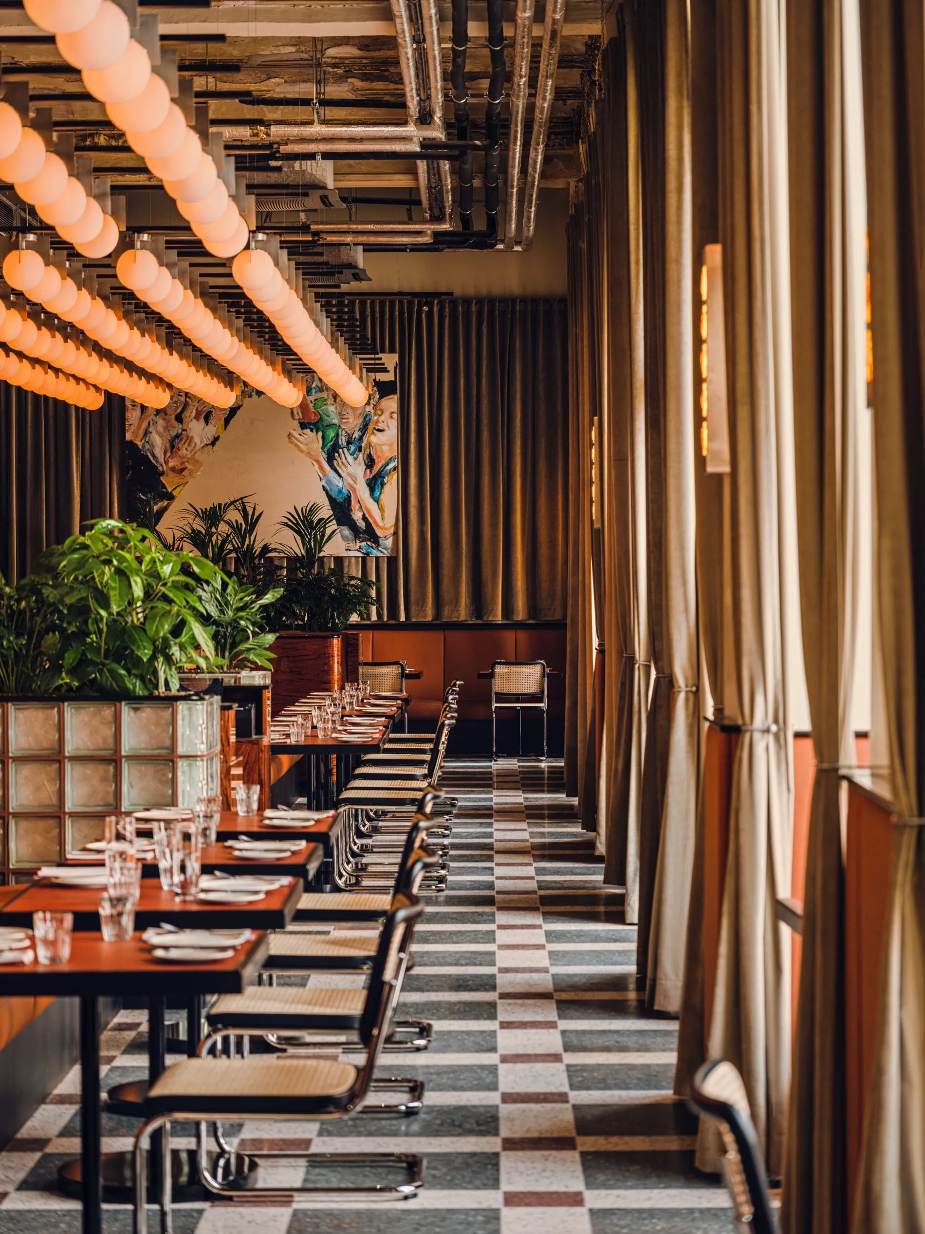 Im Restaurant Boca reihen sich eingedeckte Tische entlang bodentiefer Fenster mit Samtvorhängen. Kugelförmige Deckenleuchten und ein großformatiges Wandgemälde setzen mediterrane Akzente im industriell anmutenden Raum.