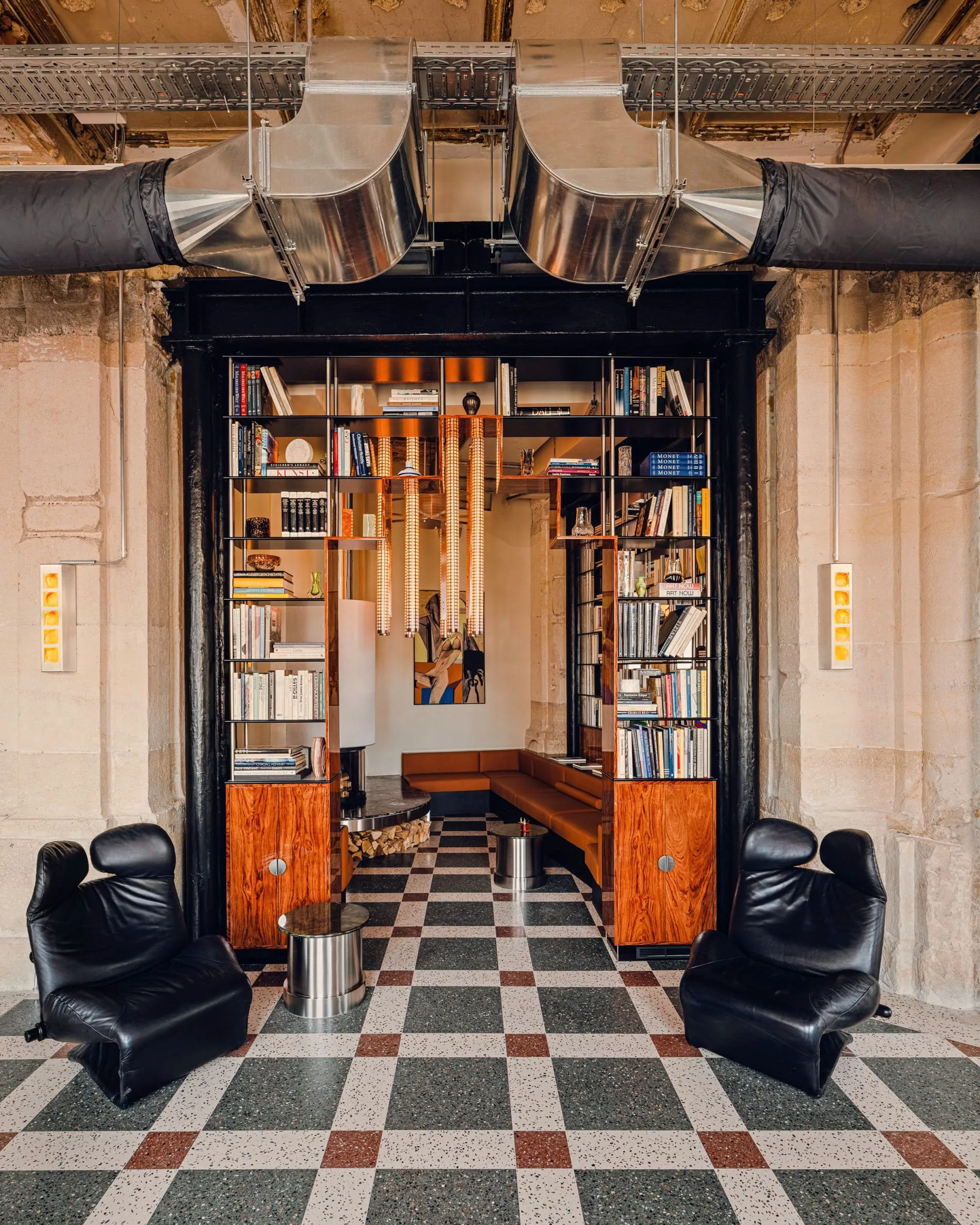 Durch einen Türbogen mit freiliegenden Lüftungsrohren öffnet sich der Blick auf Bubinga-Holzregale voller Bücher und cognacfarbene Sitzbänke. Zwei schwarze Designersessel flankieren den Durchgang.