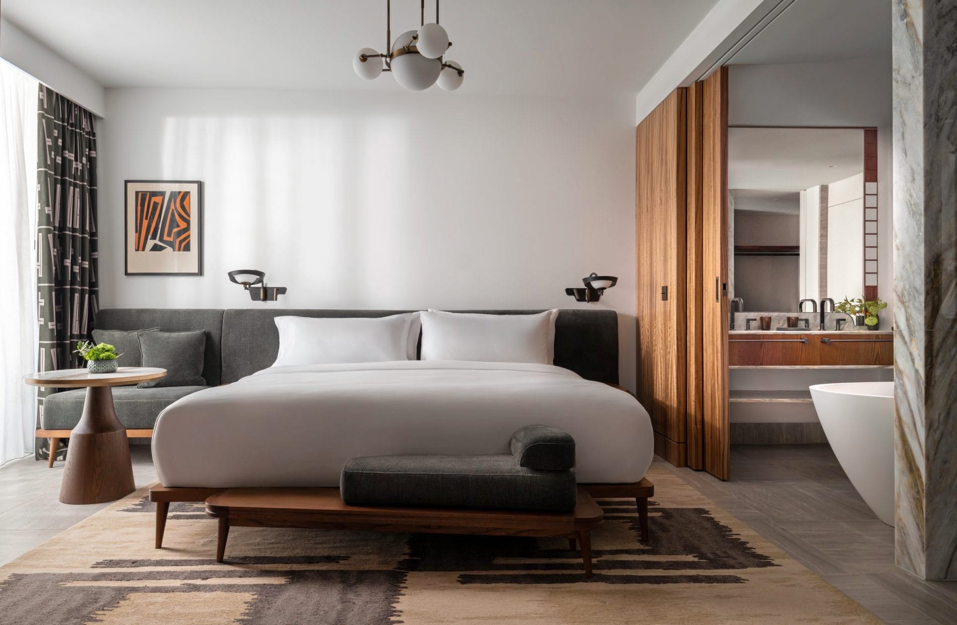 Die Deluxe Suite zählt zu den 278 Zimmern und Suiten, die laut Betreiber zu den größten Athens gehören. Mid-Century-Möbel, warme Holztöne und ein offen gestaltetes Marmorbad spiegeln die Designhandschrift des Studios Avroko wider.